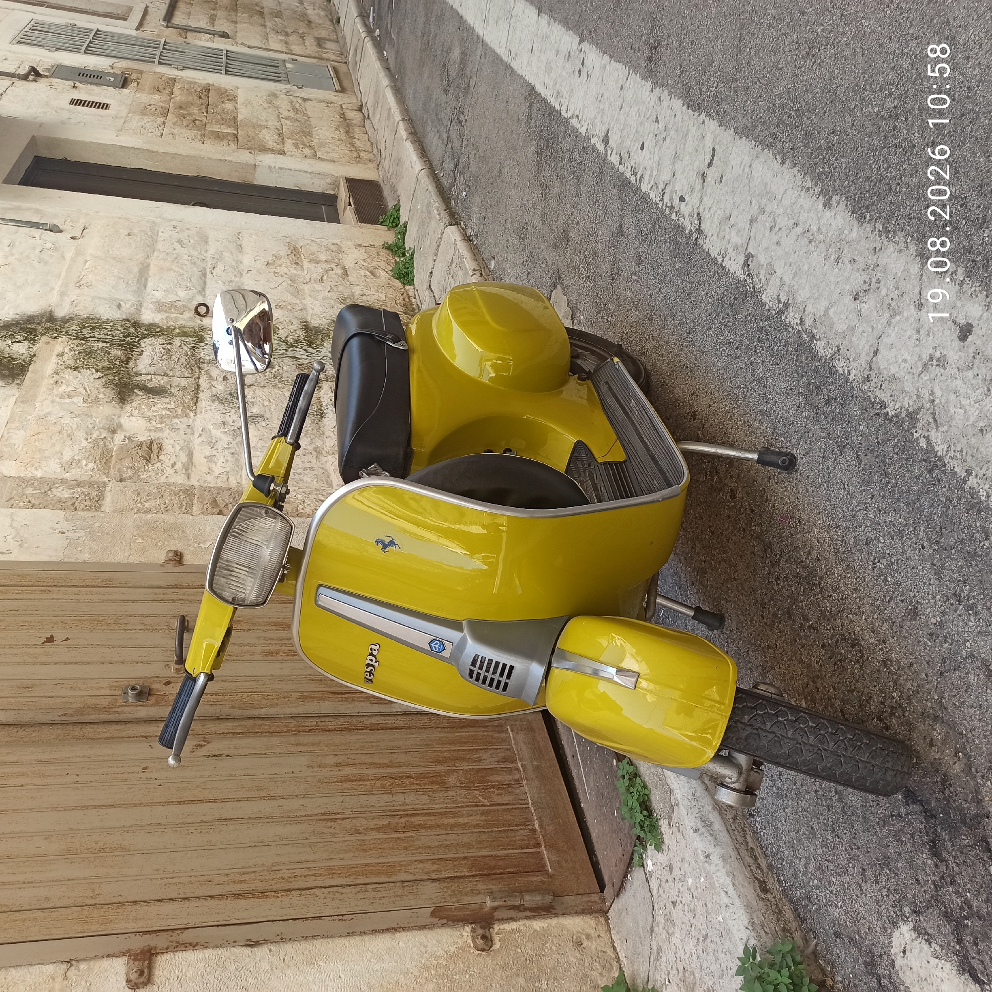 A yellow Vespa scooter is parked on a narrow street in Ruvo di Puglia, Italy. The scooter is facing slightly to the right, and its kickstand is down. The street is paved with asphalt and has a white line painted on the right side, indicating a crosswalk or lane marker. To the left of the scooter is a large, weathered wooden door set into a textured stone wall. The building to the right of the scooter also features a stone facade with a dark-colored door and a barred window. There are patches of green vegetation growing at the base of the stone wall. The sky is not visible in the frame. A timestamp "19.08.2026 10:58" is superimposed on the lower right corner of the image. There are no people present in the image.