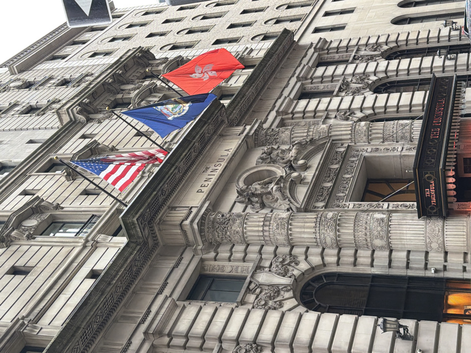 The image displays the ornate stone facade of The Peninsula hotel in New York City, United States of America, specifically identified as The Peninsula New York located at 700 Fifth Avenue at 55th Street. Three flags are mounted on flagpoles extending from the building above the main entrance: the flag of the United States of America, the blue flag of the Commonwealth of Pennsylvania, and the red flag of Hong Kong. The building exhibits elaborate Beaux-Arts architectural detailing, including sculpted classical figures above a central arched entryway, fluted pilasters, decorative cornices, and arched and rectangular windows. The name "THE PENINSULA" is carved into the stone facade and also appears on an illuminated, dark-colored canopy extending outward over what would be the street level. No people or active events are visible. The lighting is soft and even, suggesting an overcast condition. A partial white street sign with a black arrow is visible on the upper right.