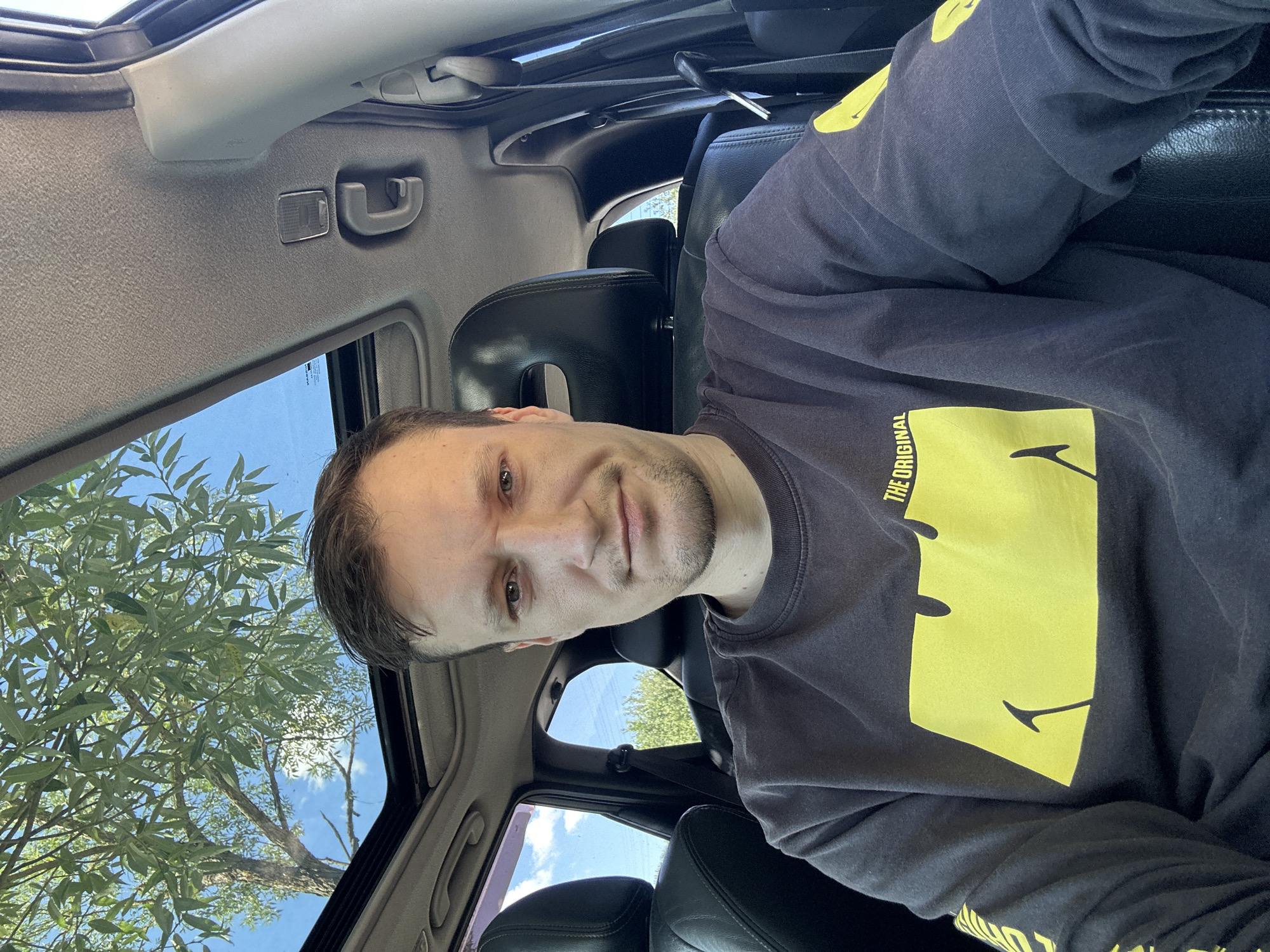 A man is sitting in the driver's seat of a car in Makiv, Ukraine. He is wearing a dark grey long-sleeved shirt with a large yellow smiley face graphic on the front and text that reads "THE ORIGINAL". He has short brown hair, a light complexion, and stubble on his chin. The car has a sunroof, which is open, revealing a clear blue sky and the branches of a green tree. The interior of the car is visible, including black leather seats and the car's headliner. The man is looking directly at the camera with a neutral expression. The sun is shining, casting light on the scene.