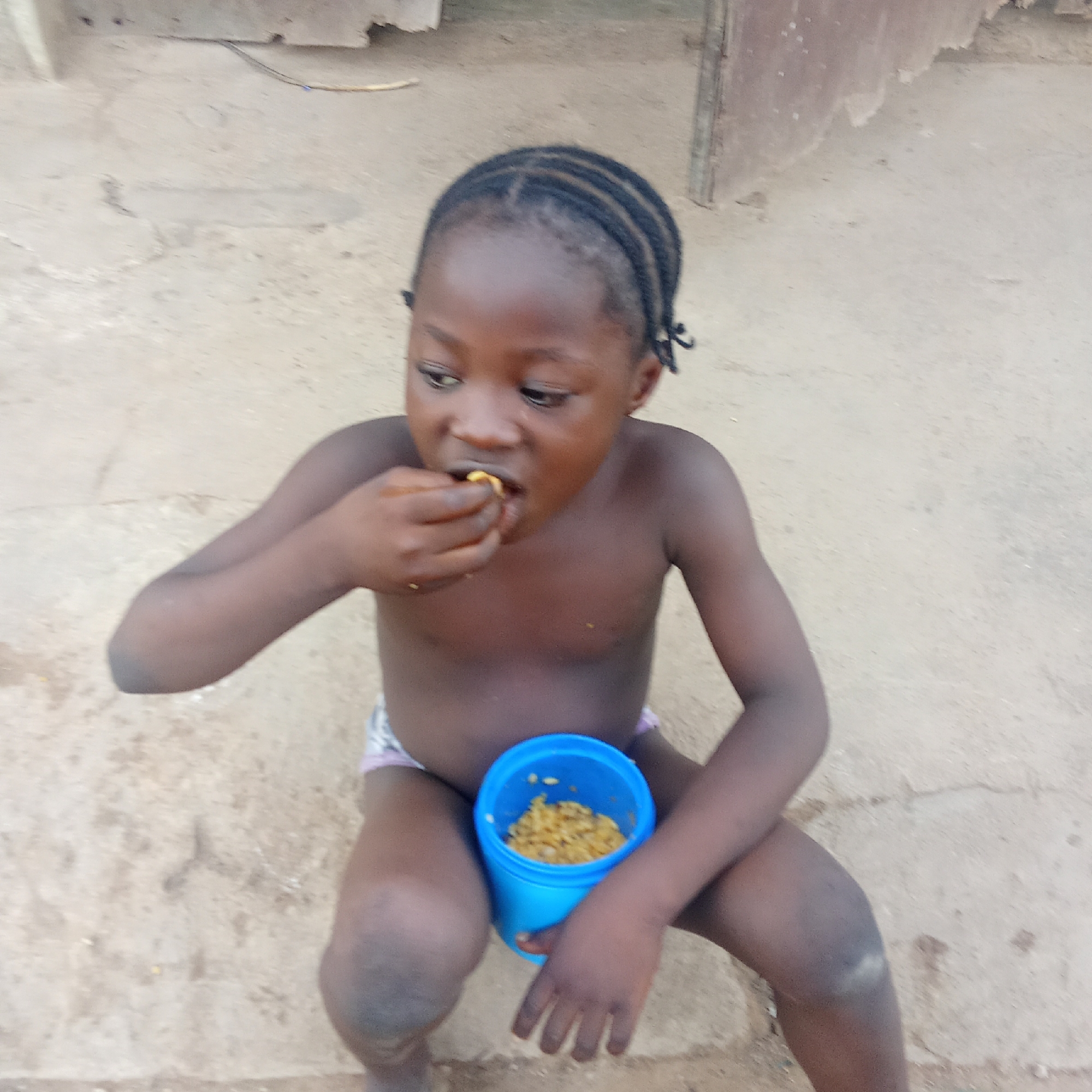The image captures a young child, likely a toddler or preschooler, squatting outdoors on a light brown, possibly dirt or concrete, ground. The child has dark skin and their hair is styled in multiple small braids. They are shirtless, wearing what appears to be light-colored shorts or underwear.
The primary activity taking place is the child eating. They are holding a bright blue cup or bowl between their legs, which contains an orangey-yellow, granular food resembling rice. The child is using their right hand to bring a small portion of the food to their mouth. Their expression is focused on eating.
Notable details include the rustic outdoor setting, with an indistinct background that suggests simple structures or an open area. The lighting indicates it is daytime. The child's posture and direct consumption of food from the cup with their hands convey a natural, everyday moment. There is no visible text in the image. The location context provided indicates this scene is in Akwanga, Nigeria.