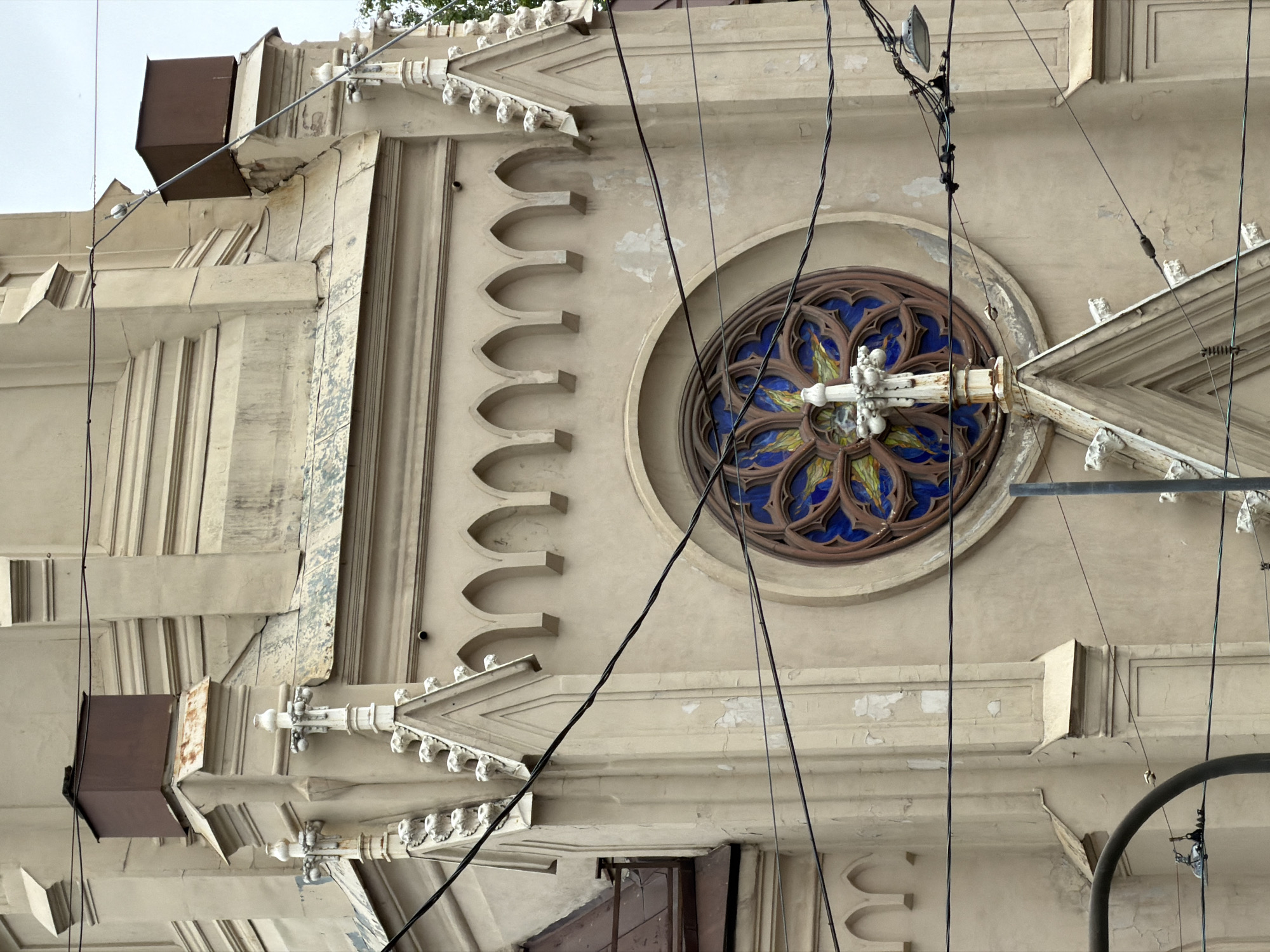 The image shows a portion of a building facade, likely a church, in Saint Petersburg, Russia. The facade is predominantly beige, and features architectural details like arched windows, decorative molding, and a large, circular stained-glass window with blue and gold panes. Electrical wires are strung across the facade, obscuring some of the details. The building shows signs of age and weathering, including peeling paint and cracks in the stonework. A dark metal box or structure is visible at the top left of the frame.