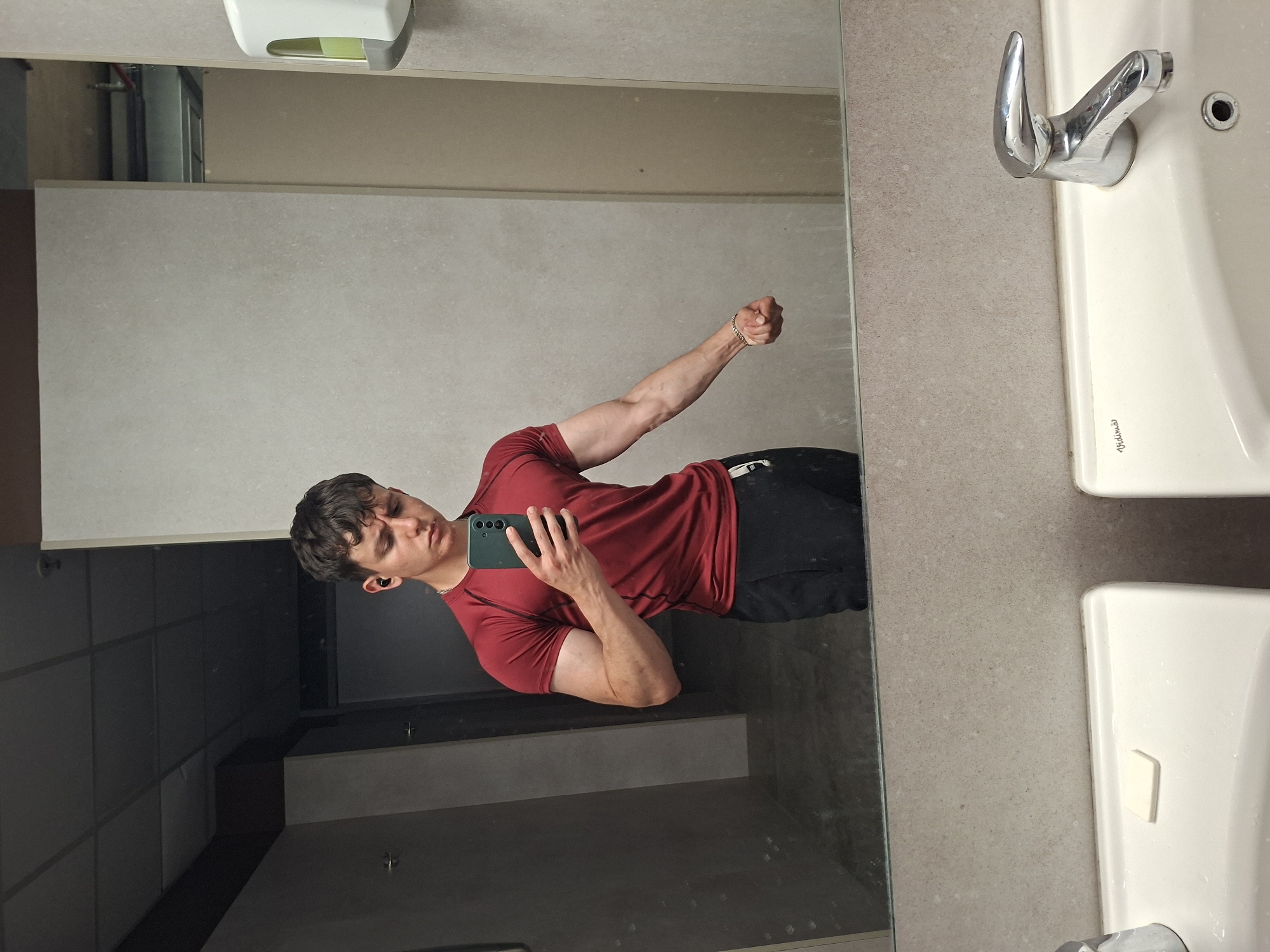 The image, rotated 90 degrees counter-clockwise, displays a young man captured in a mirror selfie. The man appears horizontally oriented in the frame due to the rotation, but his reflection shows him standing upright. He has short, dark, curly hair and is wearing a red short-sleeved t-shirt and dark trousers. He holds a dark-colored smartphone in his right hand, pointed at his reflection. His left arm is flexed, showcasing muscle definition, and he wears a gold-colored chain bracelet on his left wrist. An earbud is visible in his left ear.
The setting appears to be a public restroom or washroom, located in Plovdiv, Bulgaria. To the right of the mirror, a light grey or beige speckled countertop is visible, along with parts of two white ceramic sinks. A chrome faucet is mounted above the upper sink. Above the mirror, a white and grey liquid soap dispenser is attached to the wall. The background reflected in the mirror behind the man consists of darker, possibly tiled or paneled walls, suggesting the interior of a facility like a gym changing room or a public building.
The man is actively taking a photograph of himself in the mirror, posing with his arm flexed. His expression is neutral and focused on the camera. The word "Vidrios" is faintly visible on the ceramic basin of the upper sink.