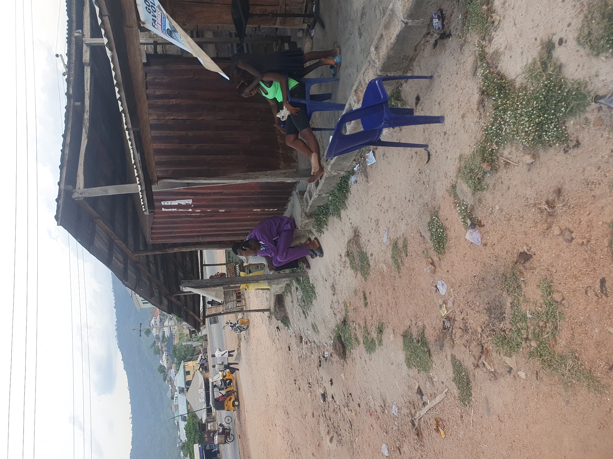 The image depicts an outdoor street scene in a town or village, characteristic of Lokoja, Nigeria. In the foreground on the left, a rustic wooden structure with a corrugated metal roof, possibly a shop or stall, is prominent. Attached to this structure is a white banner with partially visible text, including "...GOD..." and "...FAVOUR...".
Two young individuals are situated near the wooden structure. One person, wearing a bright green top and dark shorts, is seated on a blue plastic chair. Another individual in a dark top and skirt stands closely behind, possibly interacting or resting. Further along the structure, another person dressed in a distinctive purple outfit is seated on the ground or a low step.
The ground is unpaved, composed of dirt and sand, with scattered patches of green weeds and small white flowers. Litter, including plastic wrappers and paper, is visible on the ground. Beyond this, a bustling unpaved road stretches across the mid-ground, where several people are walking and congregating. Vehicles, including a yellow three-wheeled auto-rickshaw (Keke Napep) and a motorcycle, are parked nearby.
In the background, a series of buildings line the road, and towering green hills or mountains dominate the horizon. The sky is partially cloudy, suggesting daytime conditions. Power lines crisscross the upper left portion of the image. The scene conveys a casual, everyday atmosphere of a roadside community. A blurry sign in the background appears to mention "...LADY FURNITURE...".