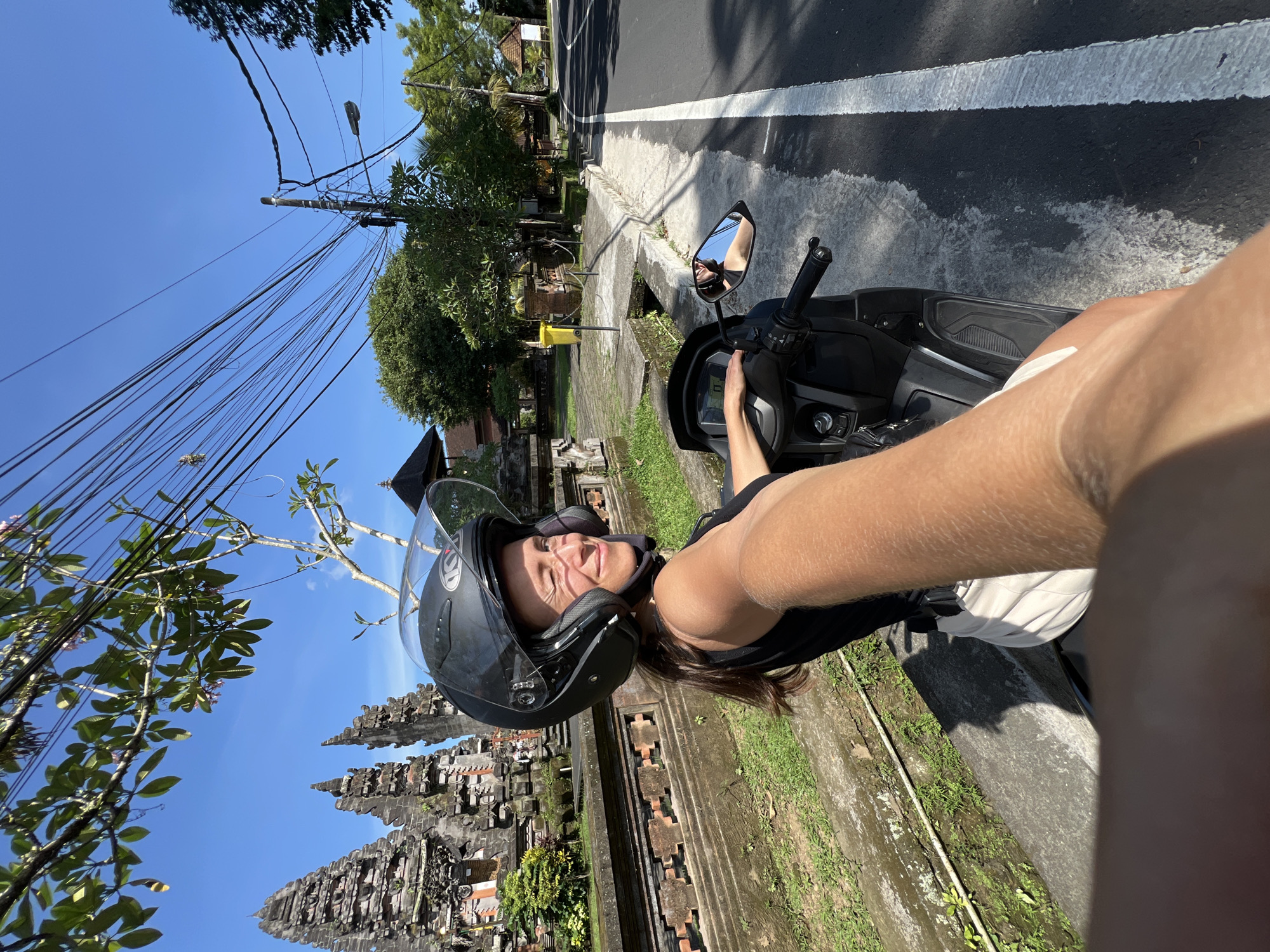 A young woman wearing a helmet is riding a scooter in Petulu, Indonesia. She is positioned on the right side of the road, next to a raised concrete curb with grass on the other side of the curb. There is a dark gray asphalt road with a solid white line marking the edge. In the background on the left is a traditional Balinese temple structure. A tangle of utility wires stretches overhead against a blue sky.