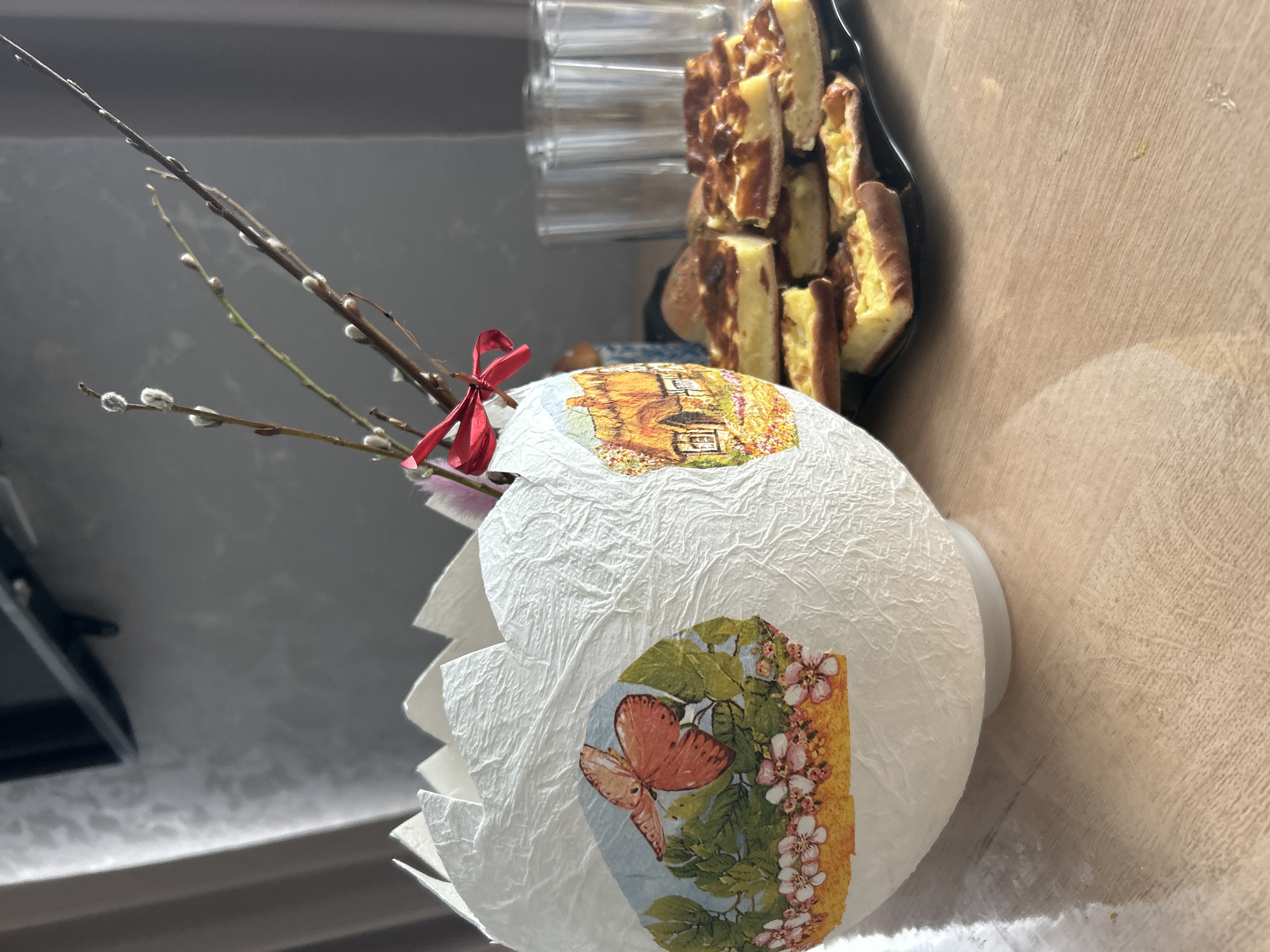 The image shows a decorated Easter egg with a butterfly and house image, adorned with pussy willow branches tied with a red ribbon, sitting on a surface next to a stack of sliced baked goods. The background suggests an interior setting, possibly a home in Chaikovsky, Russia, based on the Easter egg and the style of baked goods typically found in Russian households during Easter.