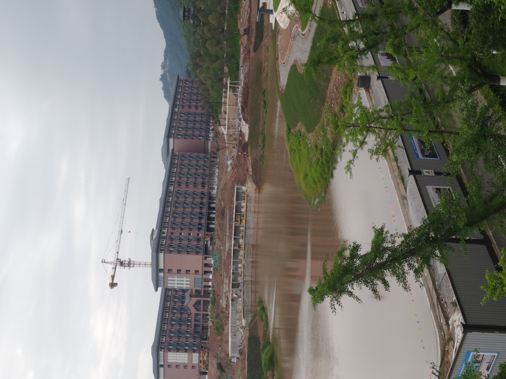 The image depicts a construction site in the Chongqing High-tech Zone, China. In the foreground, a body of murky water reflects the construction activity. Along the water's edge, there is landscaping with grass and trees. Several multi-story buildings are under construction, with a crane visible. The buildings have a brick-like facade. Construction materials and equipment are scattered throughout the site. Further away, a mountain is visible, partially obscured by clouds. Temporary structures, possibly portable offices, are visible near the water's edge. A string of colorful pennants hangs near these structures.