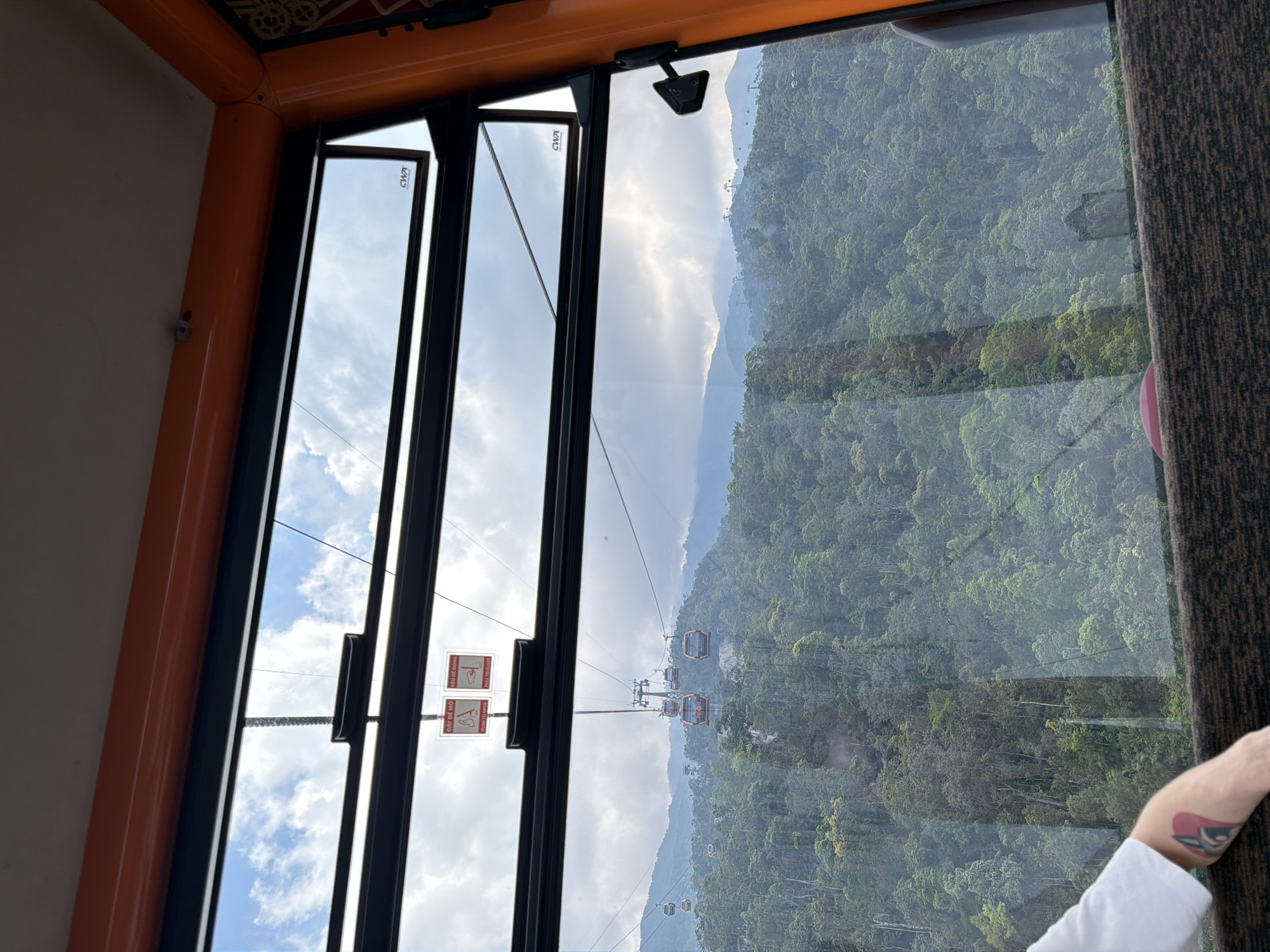 View from a cable car in Đà Nẵng, Vietnam. Lush green mountains are visible through the window. A person's arm is visible on the right side of the image. Other cable cars are visible in the distance, along the cable lines. The sky is cloudy with some sun. A safety label is affixed to the window.