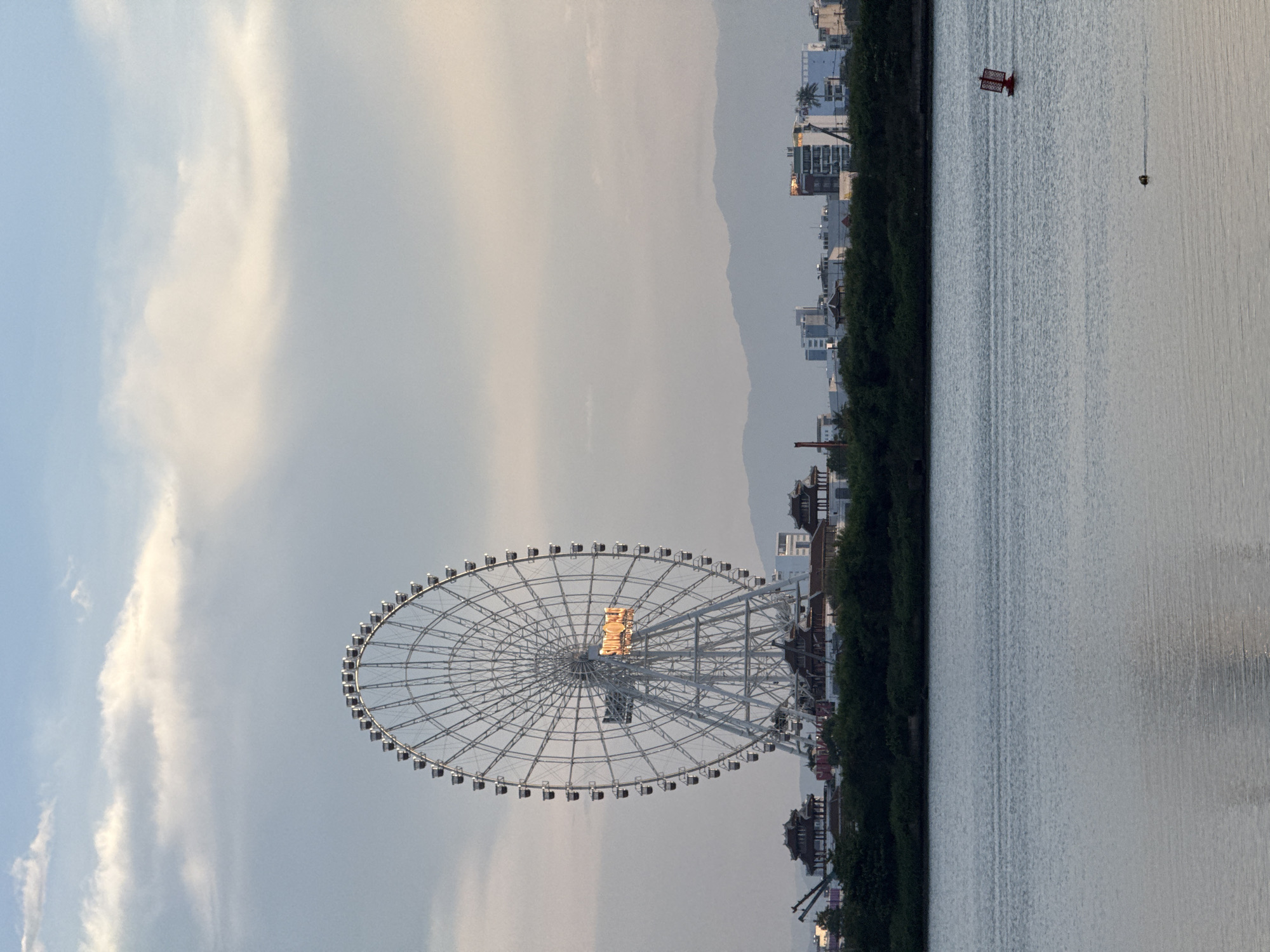 This image depicts a scenic view of the Hàn River in Đà Nẵng, Vietnam, with a prominent Ferris wheel on the left bank. The Ferris wheel is a large, white structure with numerous gondolas, some of which appear to be illuminated by the setting sun. The sun's rays reflect off the water, creating a shimmering effect.
In the background, a line of trees separates the river from a cityscape of buildings, some of which are modern high-rises, while others feature traditional East Asian architectural elements. Mountains can be seen in the far distance. The sky is partly cloudy with streaks of white clouds against a pale blue backdrop.
On the right side of the image, the river is calm, with a small red boat or marker visible in the distance. The overall atmosphere is serene and picturesque, suggesting it is late afternoon or early evening. There is no visible text in the image.