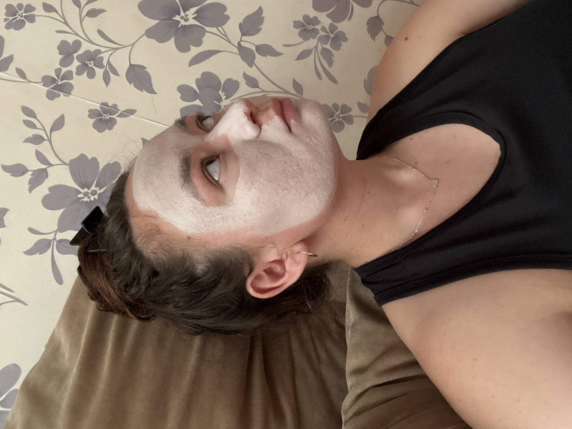 A woman lies on a brown surface, possibly a bed or couch. She is wearing a black top and a silver necklace with a small heart-shaped pendant. She has a white facial mask applied to her face. Her hair is dark and pulled back with a black clip. The background is a patterned wallpaper with gray flowers on a light background. There is no indication of the city of Chaikovsky, Russia.