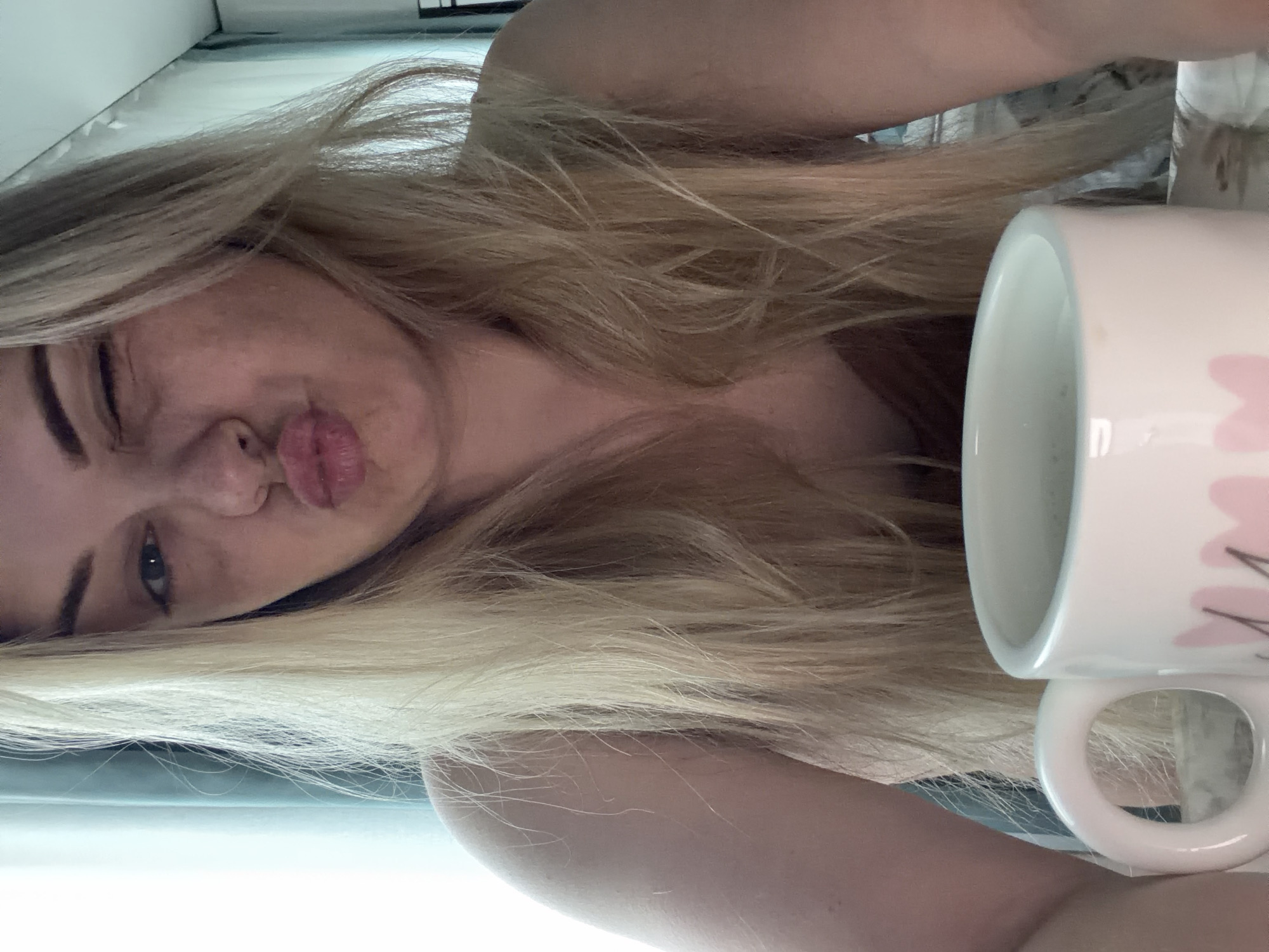 A woman with long, blonde hair is pictured close-up, her head tilted to the side. She is winking with her left eye and pursing her lips. A white mug with light pink floral designs is positioned beside her. The image is brightly lit. The location is indeterminate.