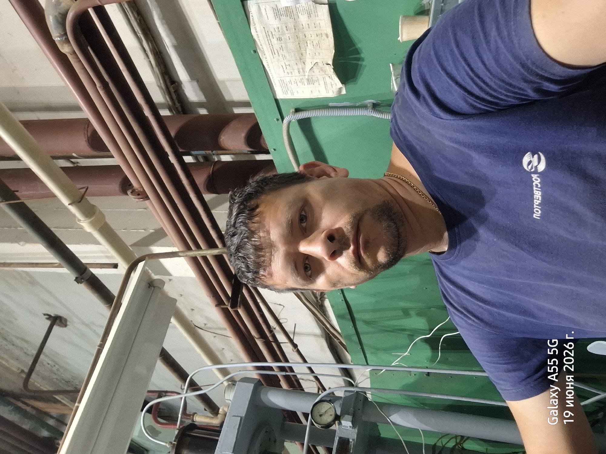 The image is a selfie of a man with short, dark hair and a goatee, wearing a dark blue t-shirt and a gold chain. He is looking directly at the camera with a neutral expression. The background appears to be an industrial setting, with various pipes, conduits, and machinery visible. A green wall or panel is present behind the man, with a document or chart attached to it. A gauge is visible on a piece of equipment to the left of the man. The bottom left corner of the image contains text indicating "Galaxy A55 5G" and a date in Russian: "19 июня 2026 г." (June 19, 2026). This suggests the photo was taken with a Samsung Galaxy A55 5G phone on that date. While the location is identified as Rostov-on-Don, Russia, the specific scene within the city is an industrial interior.
