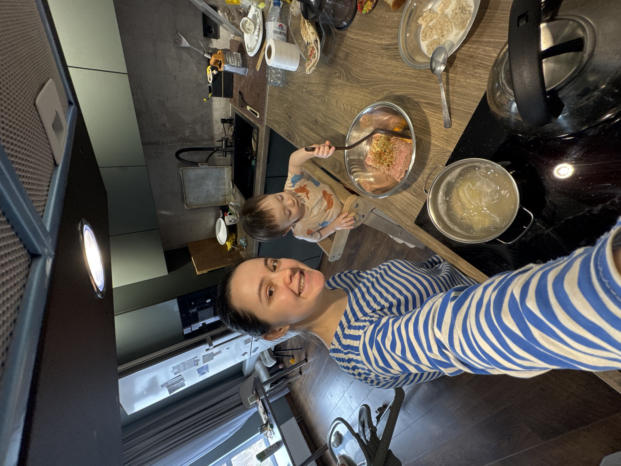 A person wearing a blue and white striped shirt is taking a selfie in a kitchen. A young child sits on a wooden high chair, stirring food in a metal bowl with a fork. A pot of boiling water is on a black stovetop next to a glass kettle. The kitchen counter also contains dishes, a blender, a box of cereal, and various cooking utensils. A sink, window, and cabinets are visible in the background. The location of the image is in Izhevsk, Russia.