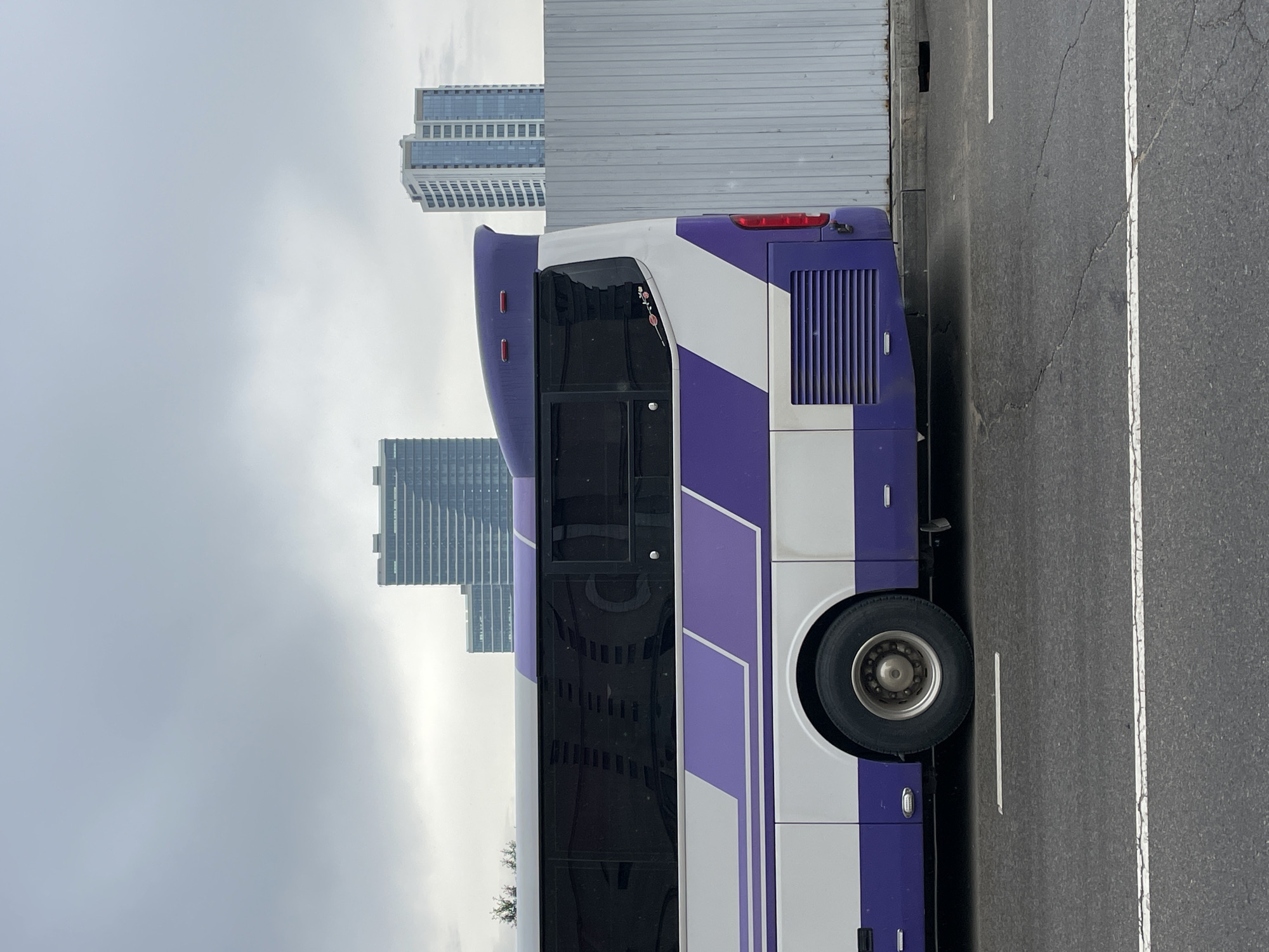 An eye-level, outdoor, full-shot photograph depicts the rear-right side of a white and purple bus parked on an asphalt road in Ulaanbaatar, Mongolia. The bus, with tinted windows and a distinctive purple spoiler, faces left, its rear bumper positioned near a light-colored corrugated metal fence. Buildings and a cloudy sky form the background. The road features white painted lane markings.