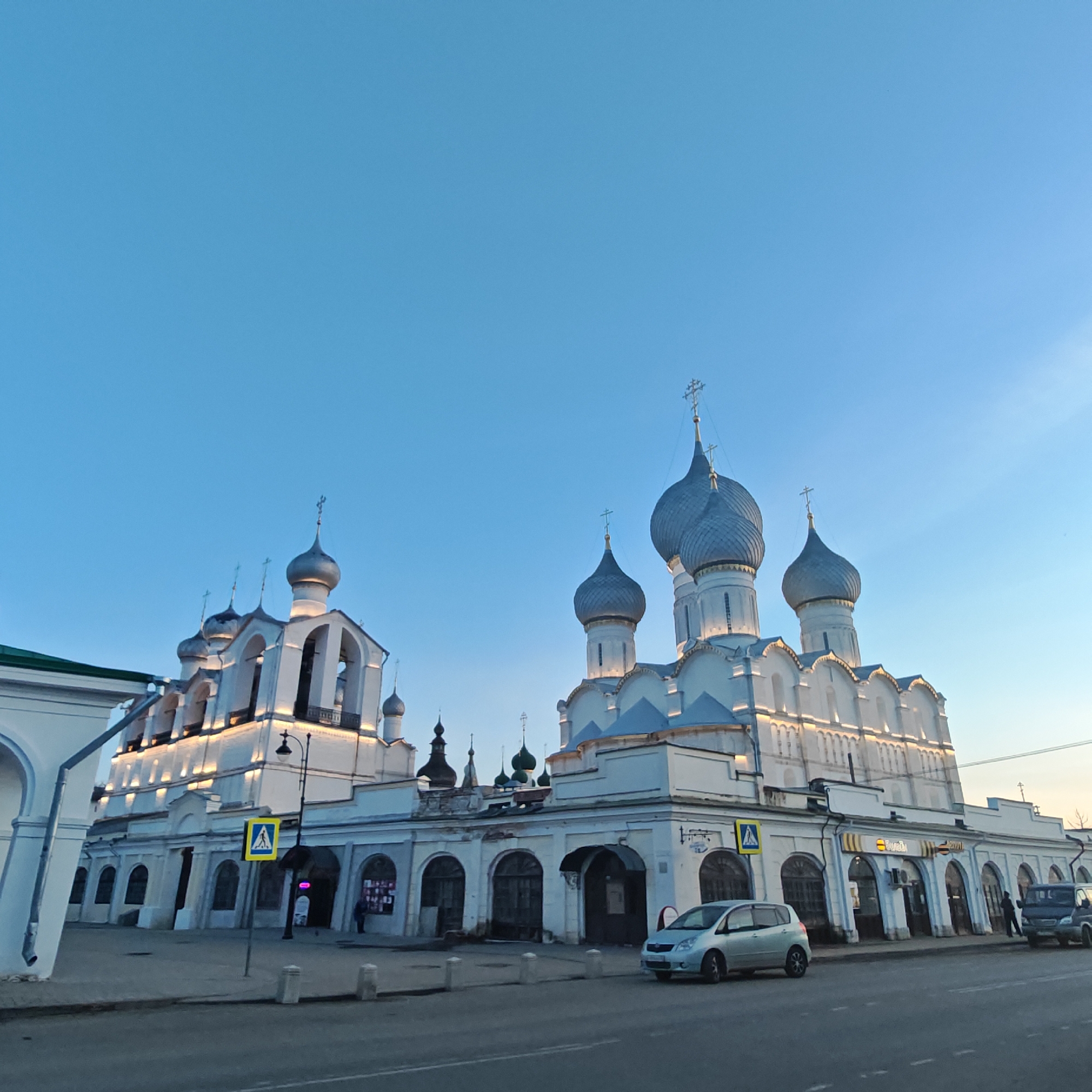 The image shows a daytime view of the city of Rostov, Russia, with a clear blue sky above. Prominently featured is a white stone building with multiple silver-domed towers, characteristic of Russian Orthodox architecture. The building appears to be a church or cathedral.
In front of the building, at street level, is a street with two parked cars and a pedestrian crossing sign. The building also includes a series of arched storefronts or entrances along its base, some of which appear to be businesses. The scene is lit with natural light, suggesting it is daytime. There are a few people walking along the storefront.