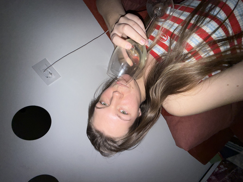 A woman is angled in the frame, holding a wine glass to her lips as if drinking. She has fair skin, light brown hair, and green eyes. She wears a red, white, and blue striped shirt. Her arm rests on a pink surface, possibly a couch. The background features a white wall with two black circular shapes and a white square with a wire protruding from it. Another wine glass is visible behind the one she is drinking from.