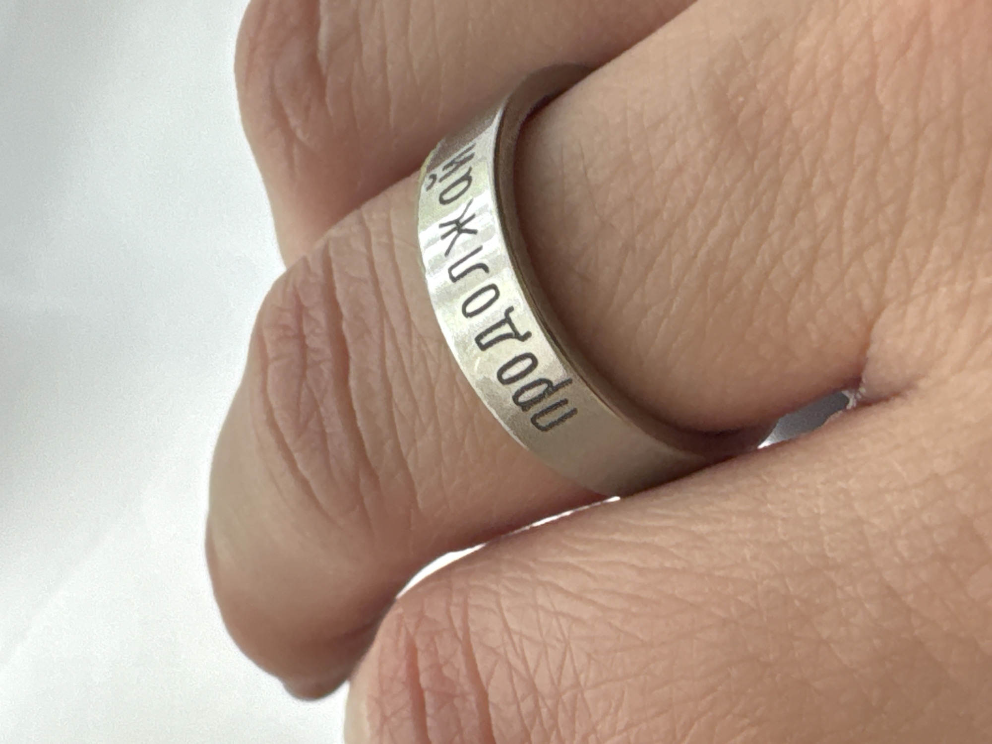 A close-up shot features a finger wearing a hammered metal ring with Cyrillic text engraved on it. The text appears to be in Russian and reads "проходи". The skin of the hand is fair and shows wrinkles, suggesting an adult. The background is out of focus and appears to be white, providing contrast and drawing attention to the ring. The ring seems to be made of silver or a similar metal. It is not possible to determine the location within Veliky Novgorod, Russia from this image.