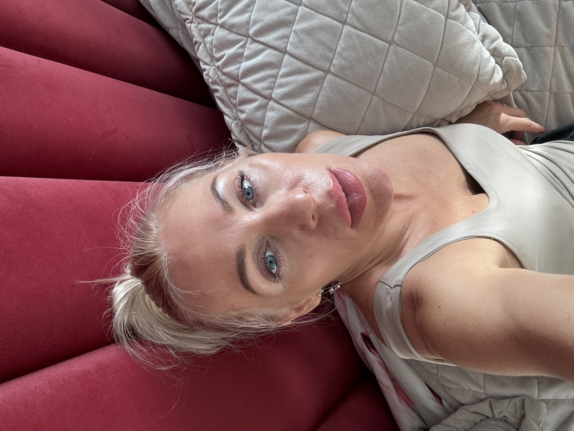 A woman with blonde hair pulled into a messy bun, bright blue eyes, and full lips looks up towards the camera. She is wearing a light-colored tank top and silver earrings. She is lying on a bed with a red tufted headboard behind her and a quilted beige blanket on the right. The lighting suggests she is indoors.