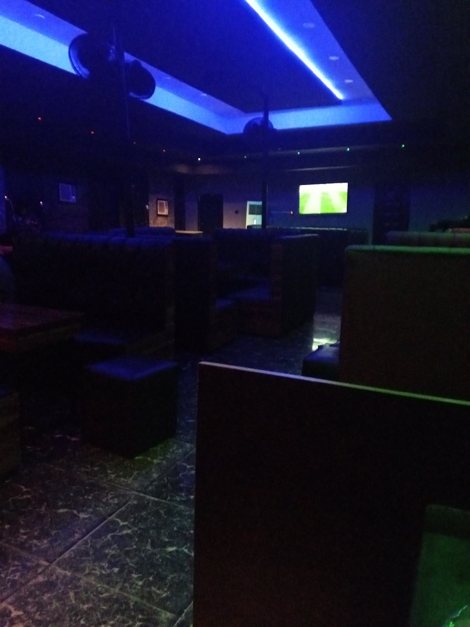 The image shows the interior of a dimly lit venue, possibly a bar or lounge, located in an unknown city and state. The lighting is primarily blue, with strips of light on the ceiling and a television screen showing a green field, likely a sporting event. The room is furnished with dark-colored booths or seating areas, separated by partitions. The floor is tiled in a dark, marble-like pattern. There are no visible people in the immediate foreground. The overall atmosphere is dark and subdued.