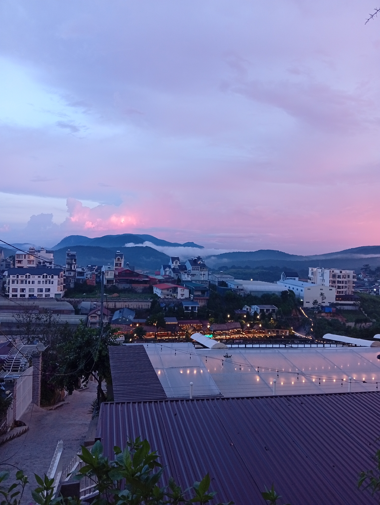 The image is a landscape-style shot of Da Lat, Vietnam at dusk. The sky transitions from pale blue to pink. Mountains are visible in the middle distance, partially obscured by low-lying fog. In the foreground, there's a view over rooftops, including corrugated metal and translucent materials with string lights. There are structures with lights, possibly a restaurant or commercial area, in the mid-ground. The city buildings are a mix of architectural styles. A road descends in the bottom left corner, near a fence. Vegetation, including leafy plants and trees, are visible in the foreground and background.