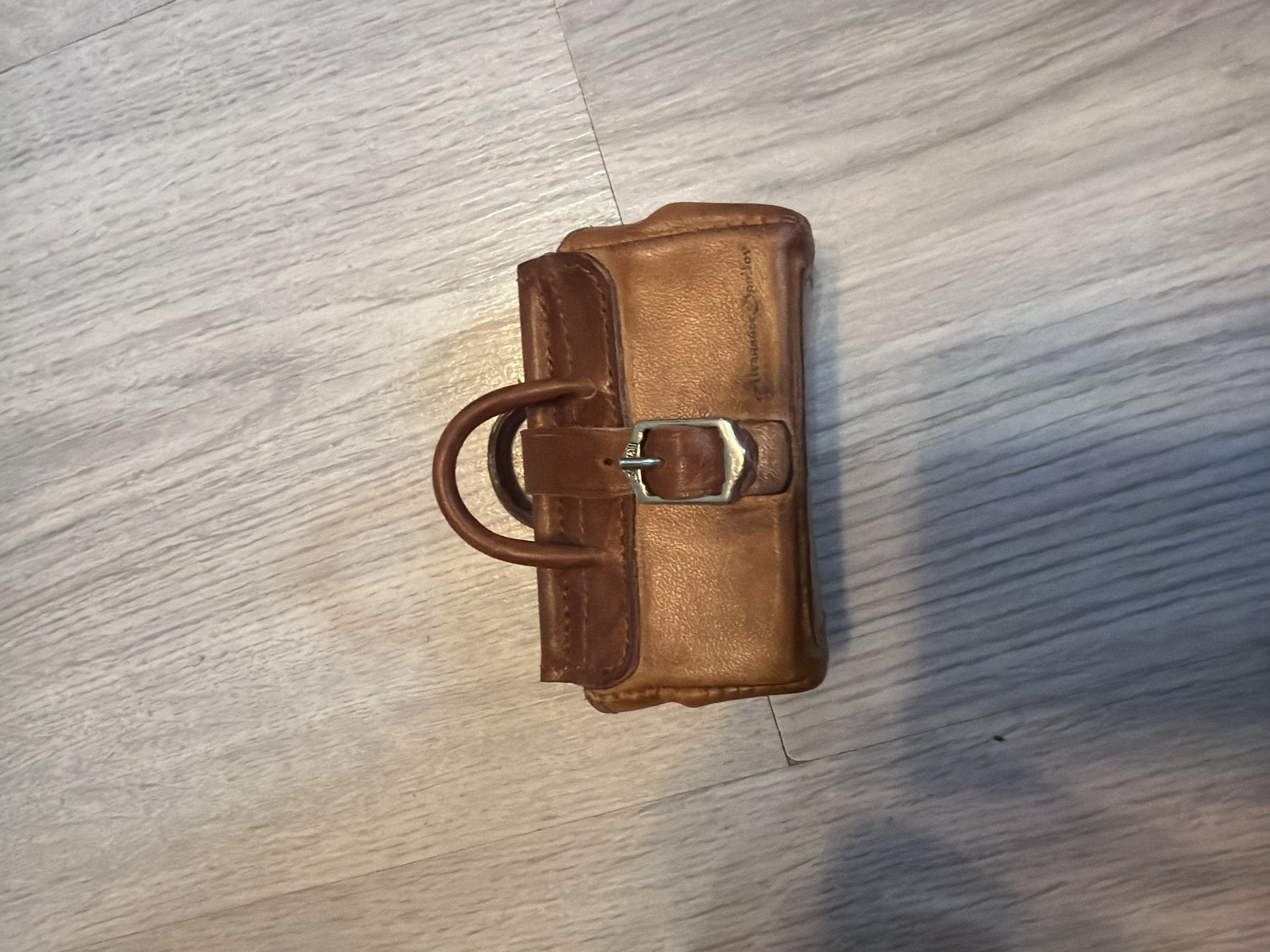 A close-up, overhead shot shows a small, brown leather handbag with a buckle and handle resting on a light-colored wooden floor. The handbag has a slightly worn appearance, with visible stitching and a subtle embossed name on the front. The wooden floor exhibits a distinct grain pattern.