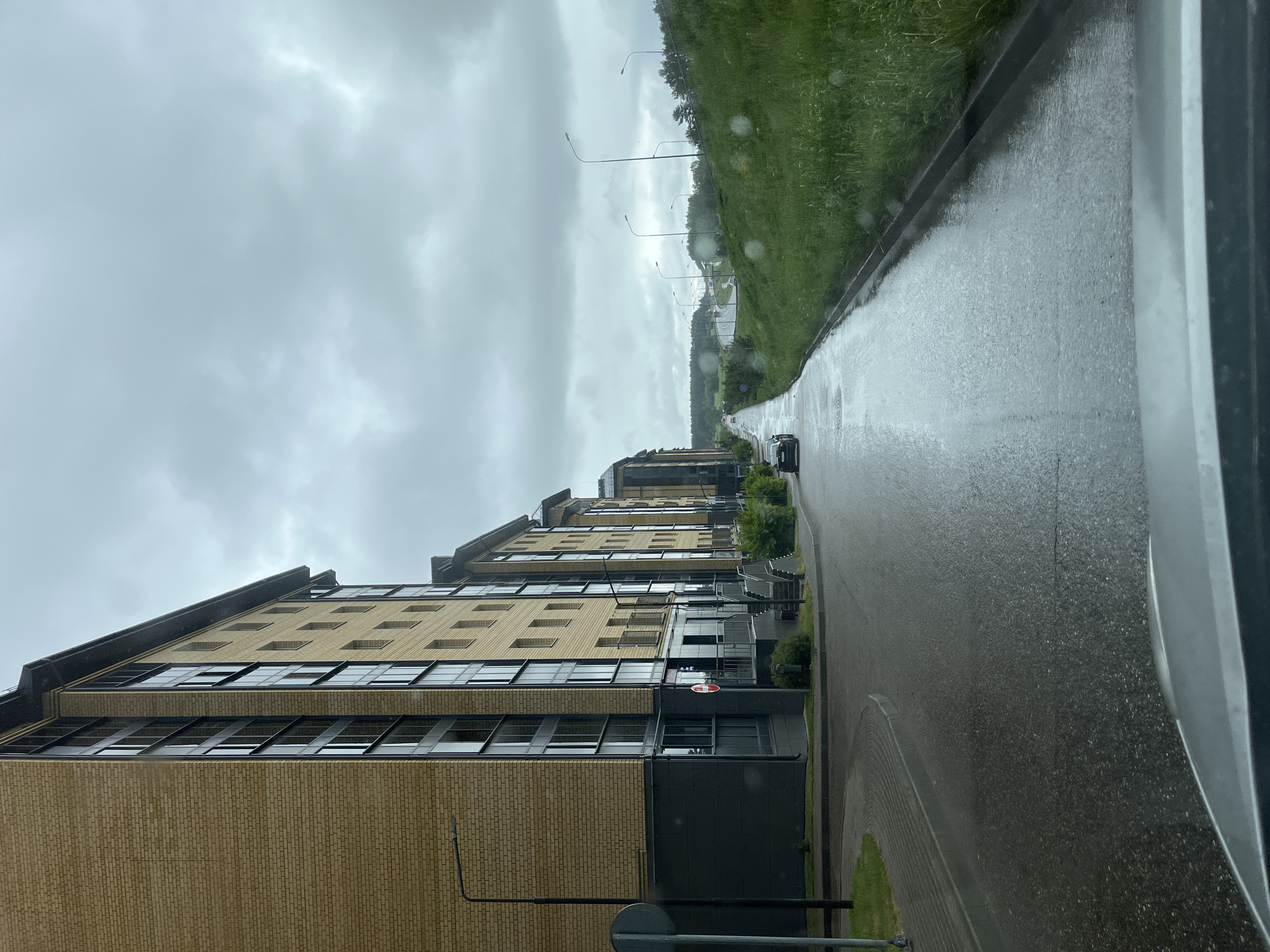 A street-level view shows a rainy day scene in Mokshino, Russia. A long, light-colored brick apartment building lines the left side of the road. The road is wet and reflects the overcast sky. A grassy hill rises on the right side of the road. Several street lamps are visible along the roadside. A car is seen driving on the road in the distance.