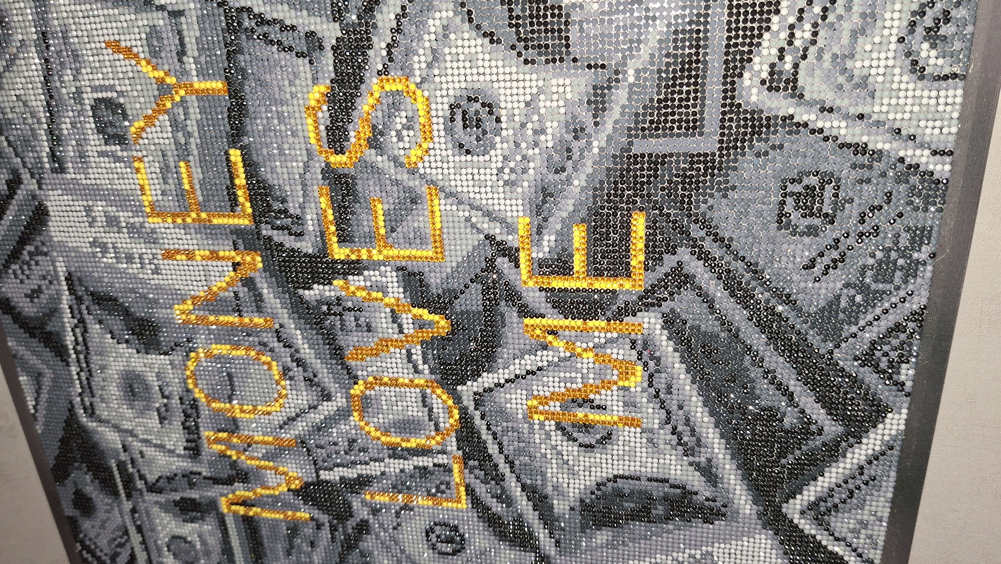The image depicts a diamond art canvas featuring a background filled with monochromatic images of U.S. dollar bills. Superimposed on the background, rendered in yellow diamonds, are the words "MONEY LOVE ME." The art piece appears to be framed. The photograph is taken at an angle, revealing the edge of the canvas and the adjacent wall. The location of the image is not evident, nor is it discernible what city or country this photograph was taken.
