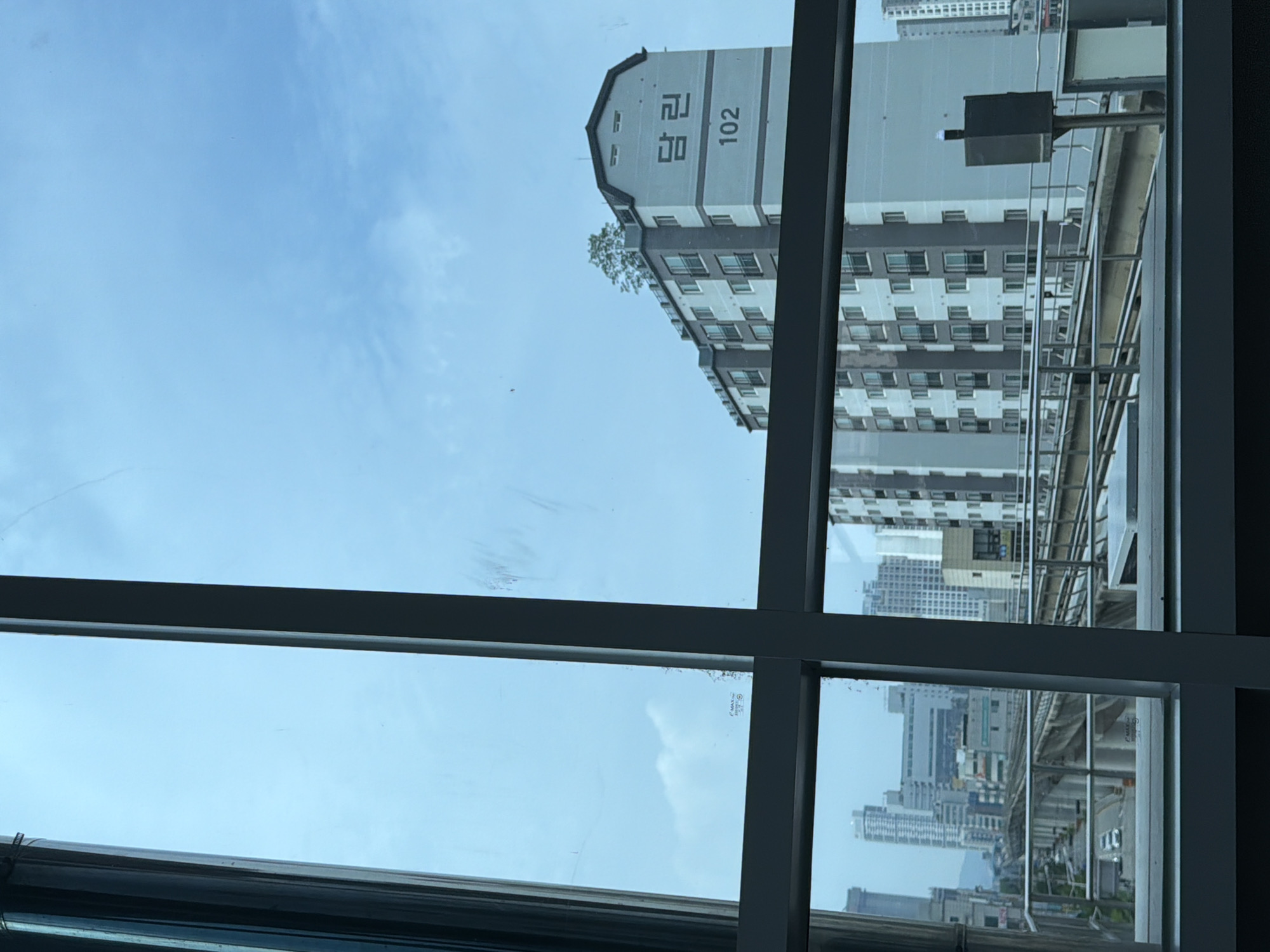 The image is a view from inside a building, looking out through a window with a grid of frames. The foreground shows a metal pillar on the left, and dark horizontal and vertical window frames.
In the mid-ground, a modern apartment building is visible. It is light gray and white, with multiple floors and many windows. The building has a peaked roof with some vegetation planted on top. On the side of the building, Korean characters that appear to read "담 린" are displayed prominently, along with the number "102".
Further in the background, a city skyline is visible, with various buildings of different heights. There is an elevated train track structure with a train or track maintenance equipment on it. Cars are visible on a road below the train tracks. The sky above is light blue with some wispy clouds. The scene appears to be in a city, possibly Uijeongbu-si, South Korea, given the architectural style and the presence of Korean characters. The overall atmosphere is urban and daytime.