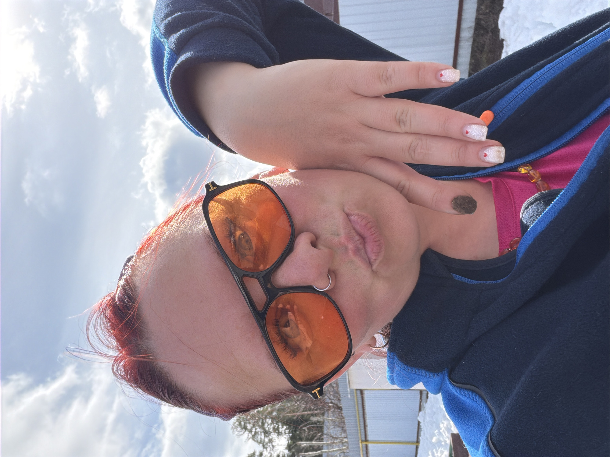 A person with red hair and orange-tinted glasses is posing for a picture in an outdoor setting, possibly in Chaikovsky, Russia. The person is wearing a black jacket with blue trim over a pink top and has their hand near their face. They have white nails with red heart accents. The location appears to be outdoors with trees, snow, and a fence visible in the background. The sky is partly cloudy.
