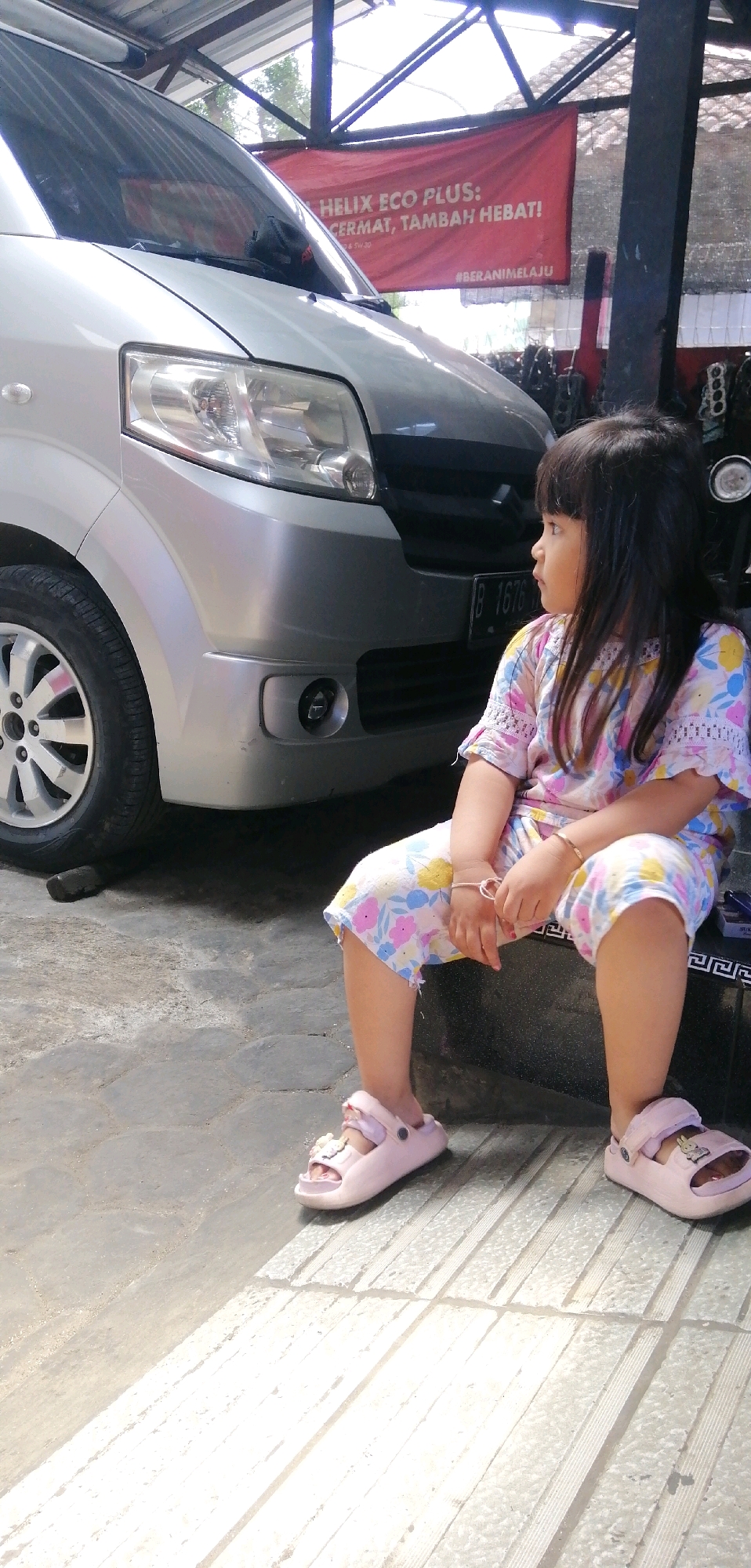A young girl with dark hair sits on a dark surface with a metallic pattern in front of a silver car. She is wearing a pink and white floral print outfit and pink sandals. Her left leg is crossed over her right leg. The car is parked under a metal roof structure. A red banner with white text is visible behind the car. The ground is paved with interlocking gray and white stones, with a visible shadow of the car extending across the foreground. The location is identified as Tinggarjaya, Indonesia.