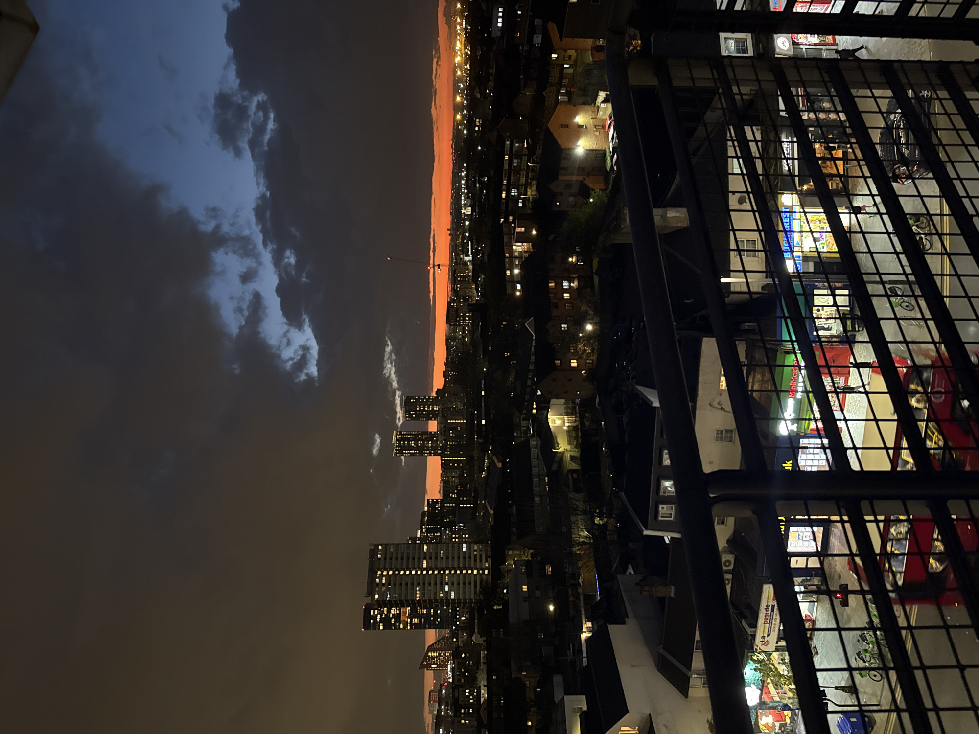 View from a high-rise balcony overlooking a city at dusk. The sky is a mix of dark blue clouds and a fiery orange horizon. Below, the city skyline includes lit buildings and apartment blocks. In the foreground, a metal railing obscures the view of the street, where a mini-market with a red car and scooters parked outside is visible. The location is London, United Kingdom.