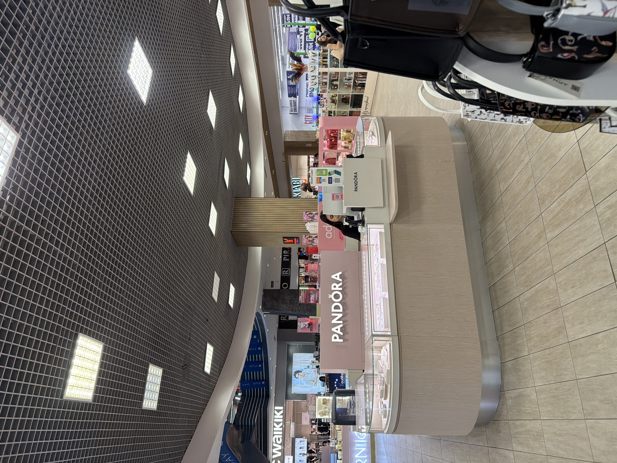 The image depicts the interior of a shopping mall. A Pandora jewelry store is prominently visible, with the brand name displayed on its pink facade. Nearby, other stores can be seen, including Waikiki and Kiabi, suggesting a variety of retail options are available. The overhead lighting consists of a grid pattern with square light fixtures. The floor is tiled in neutral tones. The scene appears to be located in Yerevan, Armenia, based on visible Armenian text on some of the store signs.