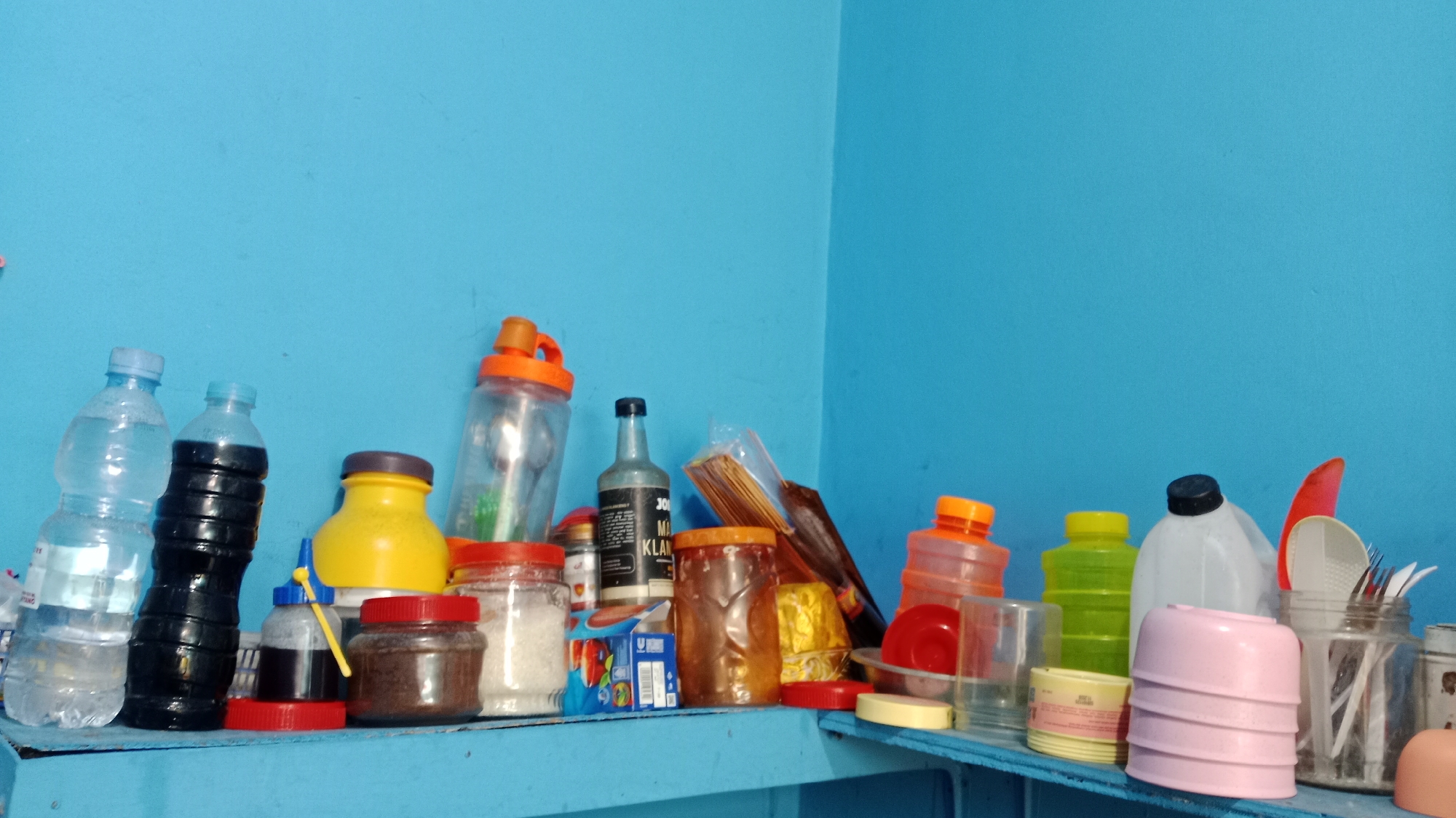The image shows a shelf against a blue wall. On the shelf are various items, including plastic bottles, jars, and containers, some filled with liquids or solids. To the left, there are two plastic water bottles, one clear and one black. Next to them is a yellow container with a dark lid, and a smaller jar with a blue cap and a yellow dispenser. Further right, there is a clear plastic bottle with an orange lid and a spoon inside. There are also jars containing white and brown substances, a bottle of dark liquid, and some packaged items. In the center and right of the image, more containers are visible, including a green bottle, a white jug with a black lid, and several pink stacked items. A red object, possibly a scoop or lid, is also present. A glass jar filled with utensils is on the far right. The overall impression is of a shelf storing food items, kitchen supplies, or cleaning products. The scene is set indoors, likely in a home or small shop, given the collection of common household items and the simple blue wall. The specific location in Denpasar, Indonesia, cannot be determined from the visual information alone.