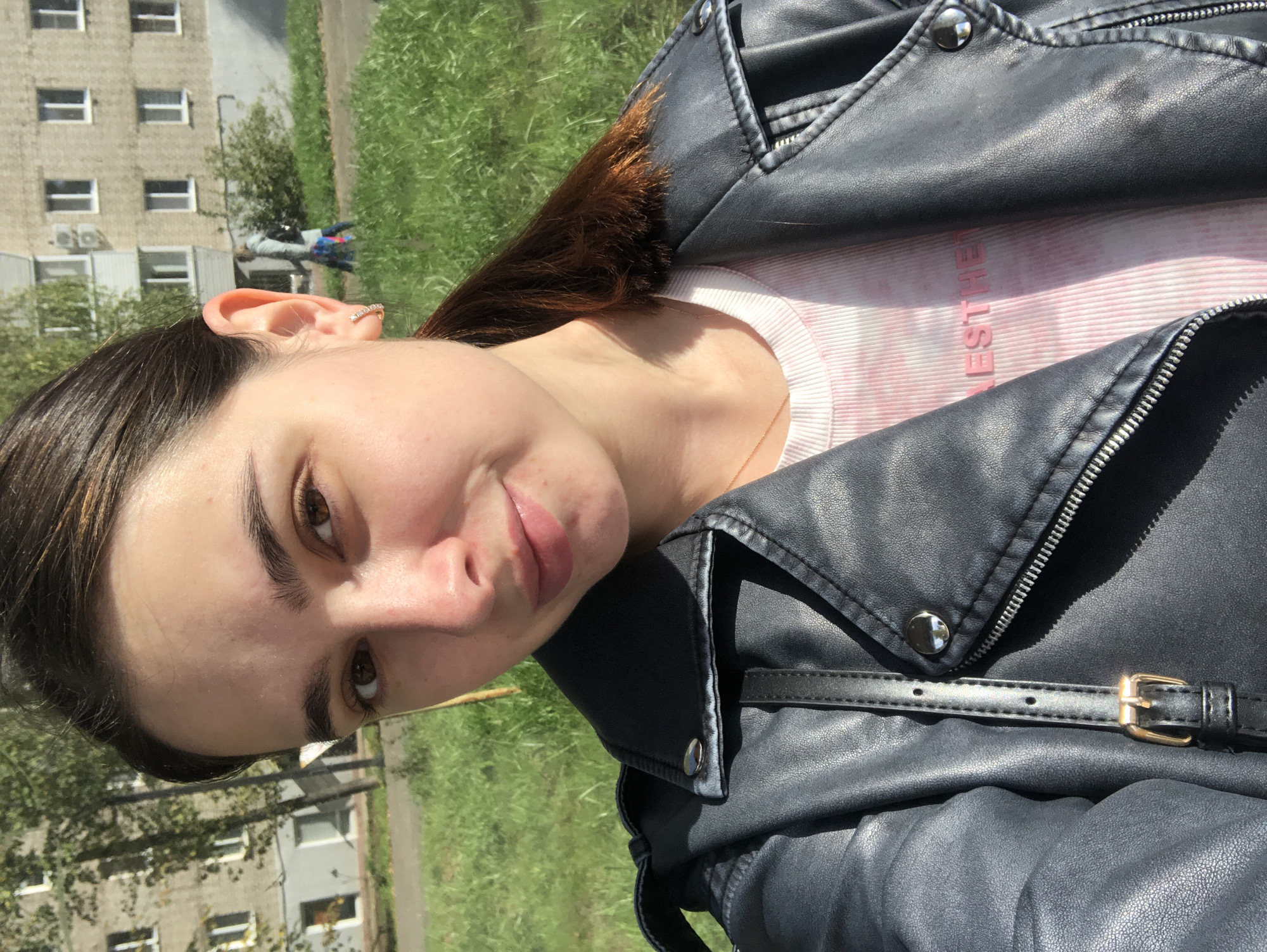 A young woman with brown hair pulled back in a ponytail and brown eyes is taking a selfie outdoors. She is wearing a dark gray faux leather jacket over a pink and white tie-dye t-shirt. A delicate gold necklace is visible. The background shows a building with a light gray facade and white-framed windows, likely an apartment building, along with green grass and trees. The scene appears to be set in a residential area of Chaikovsky, Russia, judging by the architectural style of the building. The lighting suggests it is daytime, with sunlight casting shadows. In the background, another person is walking away from the camera.