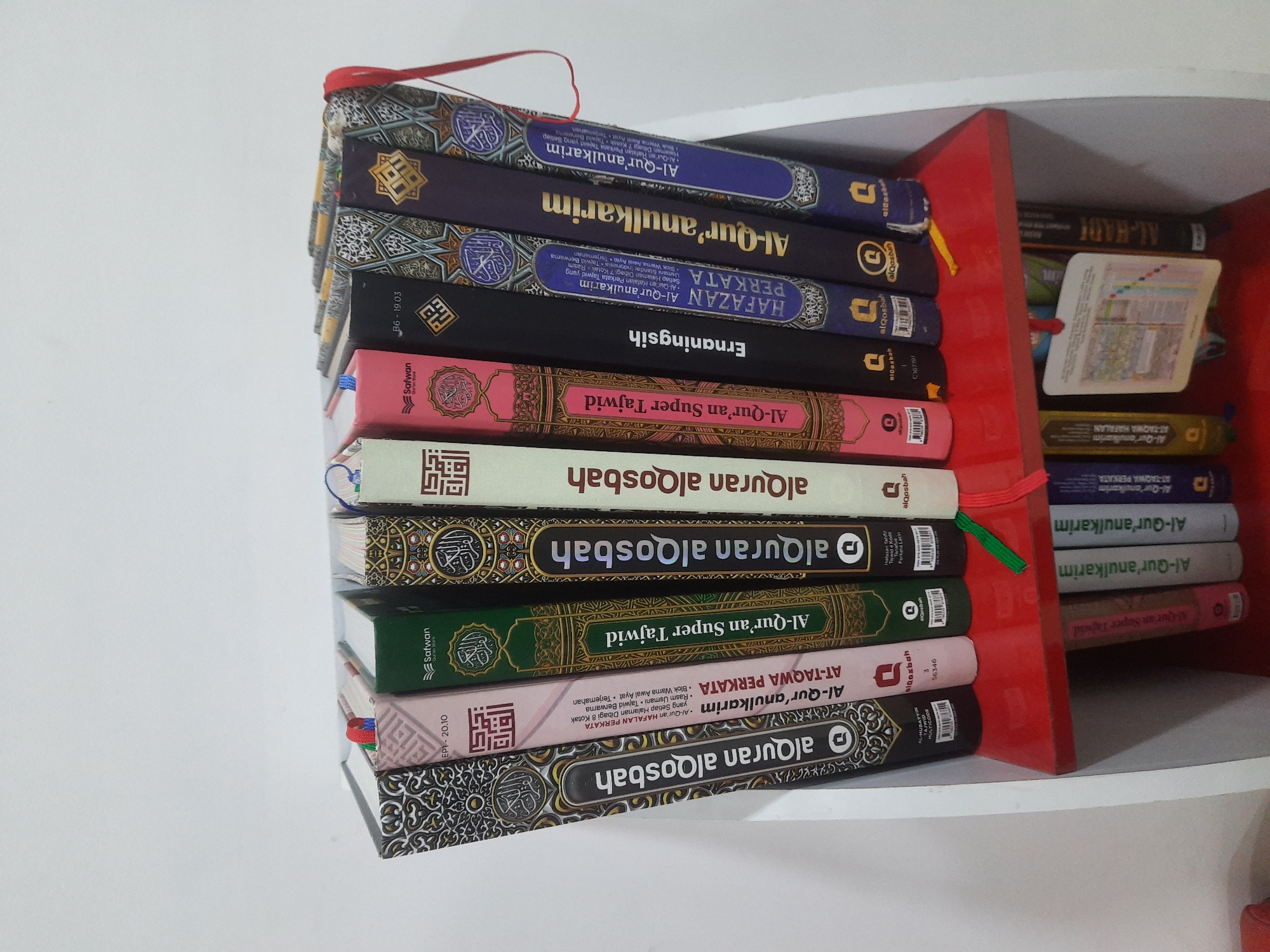 An eye-level, medium shot captures a white bookshelf filled with Islamic books. The bookshelf is predominantly white with a bright red shelf holding a selection of books. The books are arranged in a row, standing upright and leaning slightly against each other. Some books are larger and have decorative covers, while others are smaller and have simpler designs. The spines of the books display titles in Arabic and other scripts. The lighting in the scene is bright, casting minimal shadows. The background is a plain white wall, indicating an indoor setting. The overall impression is one of an organized collection of religious texts. The image does not contain any people.
