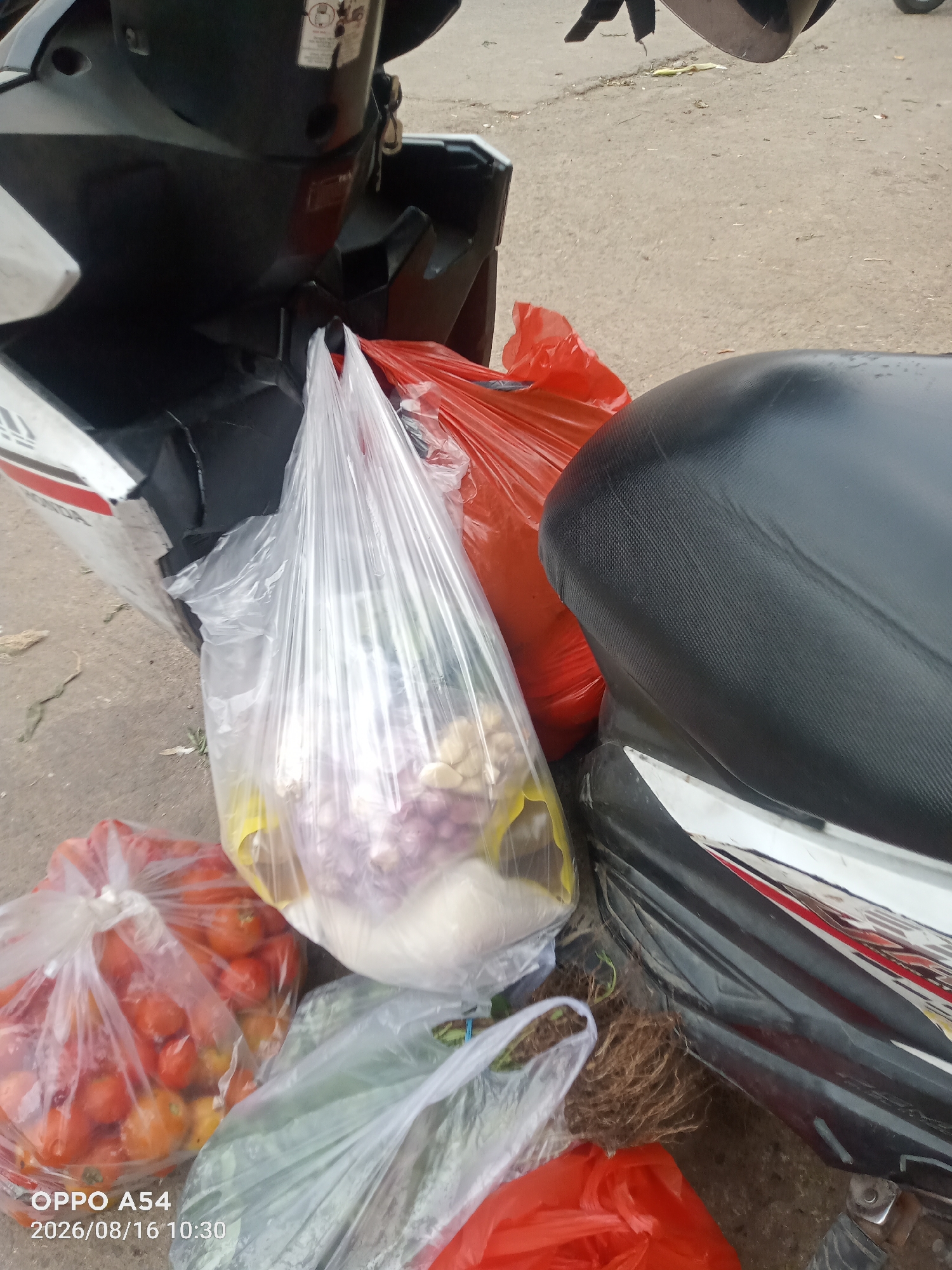 An overhead shot shows various groceries, including a bag of tomatoes, a bag of onions and garlic, and other vegetables, placed on the ground next to a parked motorcycle. The motorcycle appears to be black and white with red accents, and its seat is visible. The ground is a paved surface, likely a street or sidewalk, with some debris scattered around. The image is dated August 16, 2026, at 10:30 AM, and the watermark "OPPO A54" suggests the photo was taken with that phone model. The location is specified as Jambi, Indonesia.