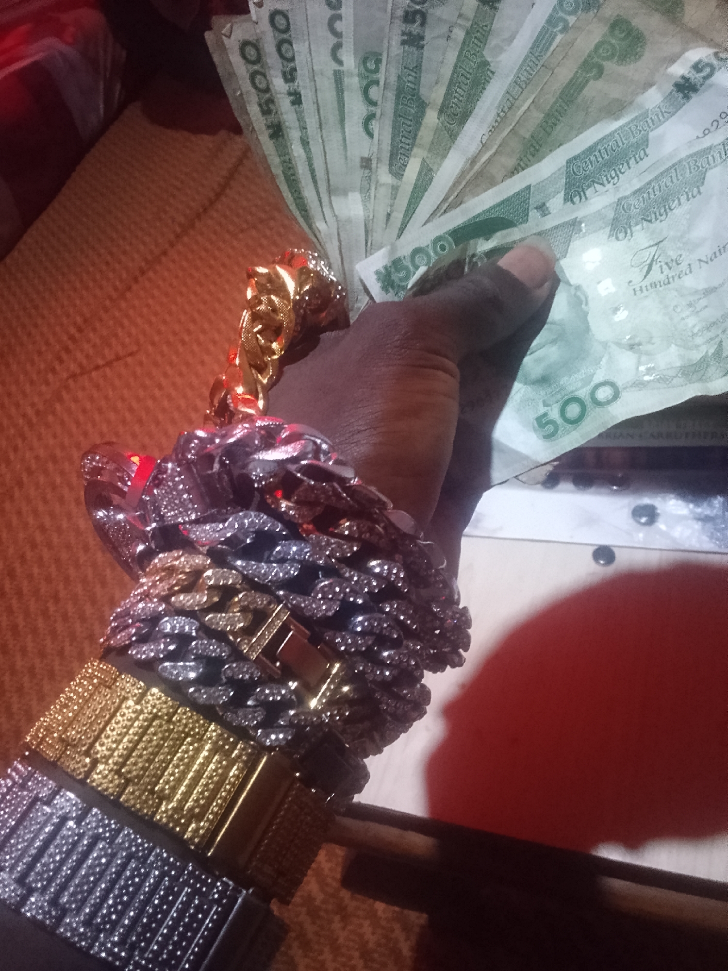 A dark-skinned person is holding a stack of Nigerian 500 Naira notes. The person is wearing multiple bracelets and a gold necklace. The scene appears to be indoors with an orange-colored background. The location and specific city are unknown.