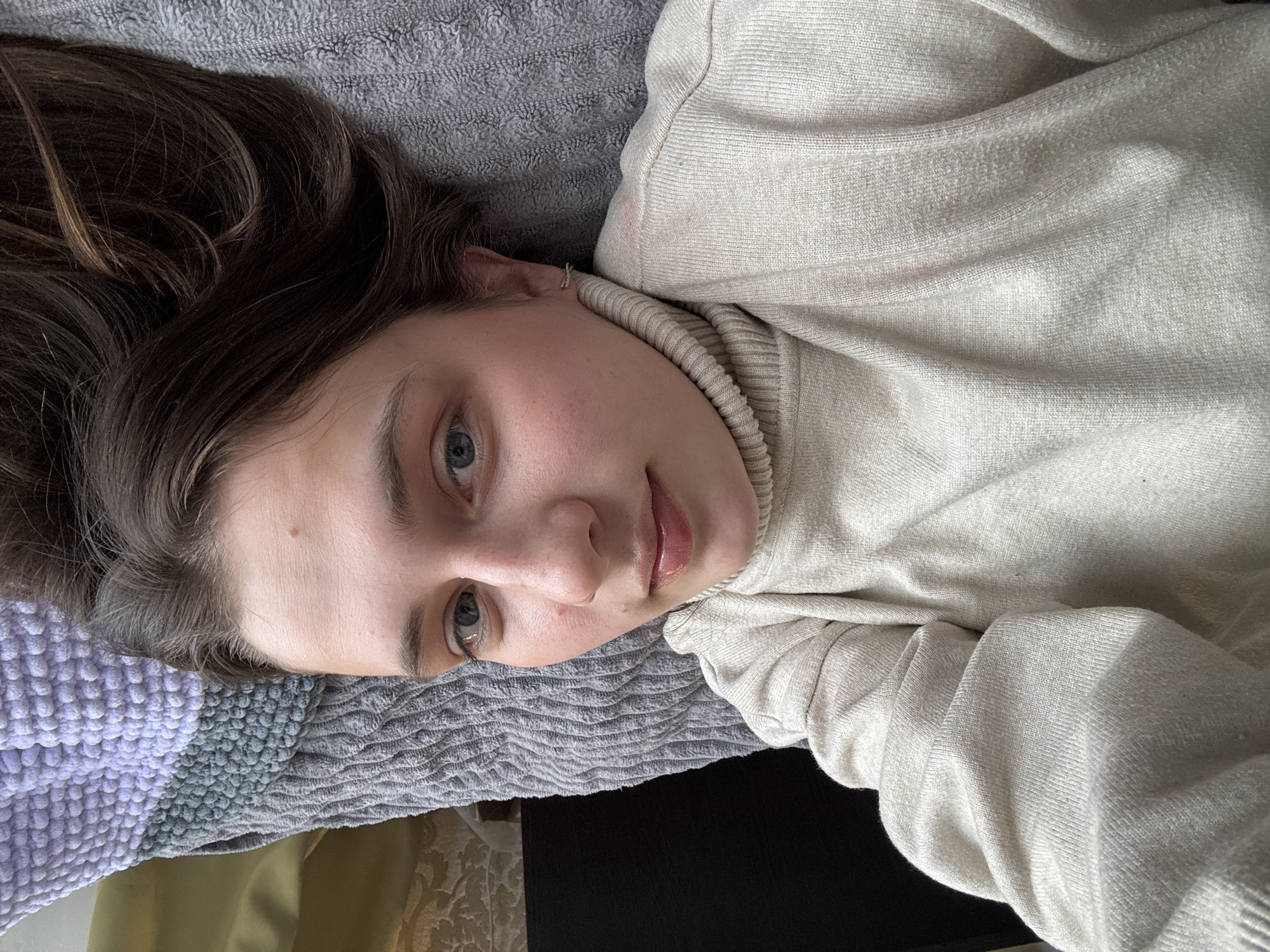 A young woman with brown hair and blue eyes is lying down, possibly on a bed or sofa, with her head resting on a patterned gray and purple blanket. She is wearing a light beige turtleneck sweater. The background includes glimpses of olive green and black, possibly furniture or other bedding. The location is unspecified, there is no indication this image was taken in the city of Chaikovsky, Russia.