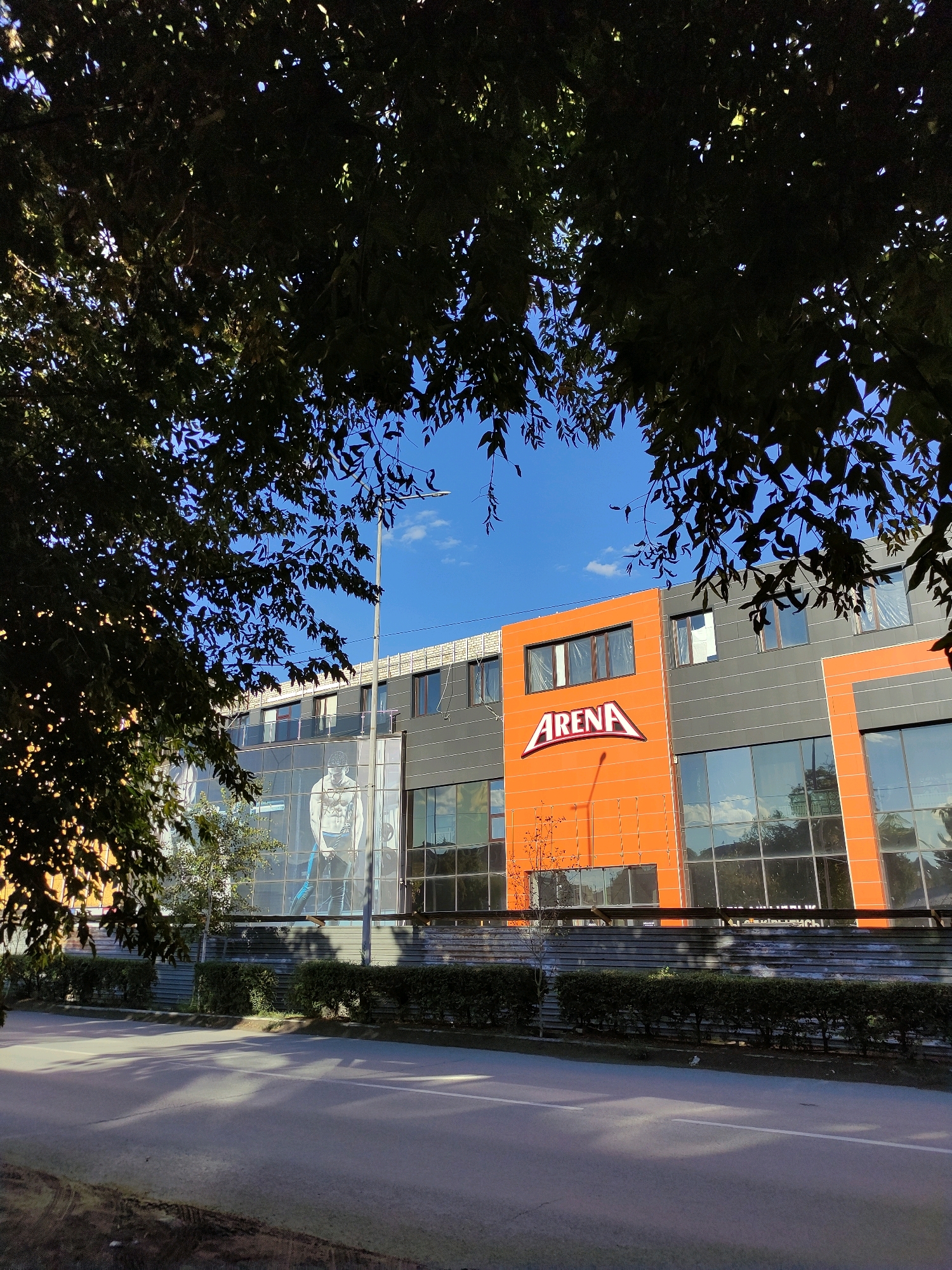 The image depicts a modern building with an orange facade featuring the word "ARENA" in large white letters. The building has large windows and is surrounded by a street and greenery. A streetlight is visible on the right side of the building. Trees with dark green leaves are in the foreground, partially obscuring the view of the building. The sky is blue with a few clouds. The image appears to be taken during the daytime, with sunlight casting shadows on the street. The overall impression is one of a commercial or recreational facility in an urban setting.