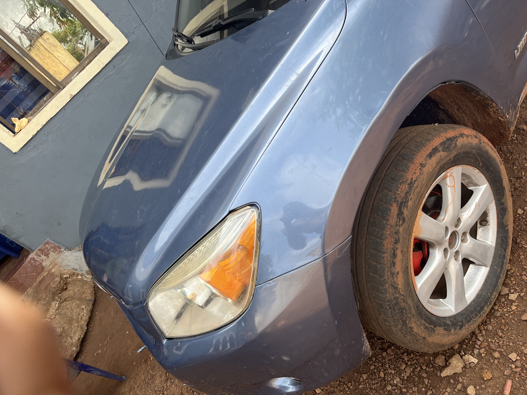 A close-up, high-angle outdoor shot captures the front passenger side of a blue car, likely a Toyota given the faintly visible lettering on the fender. The vehicle appears dusty and has minor scratches on its paintwork. The front right headlight, featuring a clear lens and an orange turn signal, is prominent. The tire beneath it is heavily caked with reddish-brown dirt, contrasting with the silver-colored wheel rim; a red brake caliper is visible behind the spokes.
The car is parked on unpaved ground consisting of reddish-brown dirt and small stones, indicating a rural or unpaved urban setting in Idah, Nigeria. Next to the car is a building with a dark grey wall. A window on this wall reflects green foliage and parts of the surrounding environment, with a small, light-colored object resting on its sill. The car's hood also shows reflections of the environment.
The scene is illuminated by bright daylight, suggesting late morning or early afternoon. A partially visible human hand or arm in the lower-left corner indicates the presence of a photographer. The overall condition of the car and the ground suggests it has been driven on dusty or unpaved roads, consistent with a vehicle in active daily use.