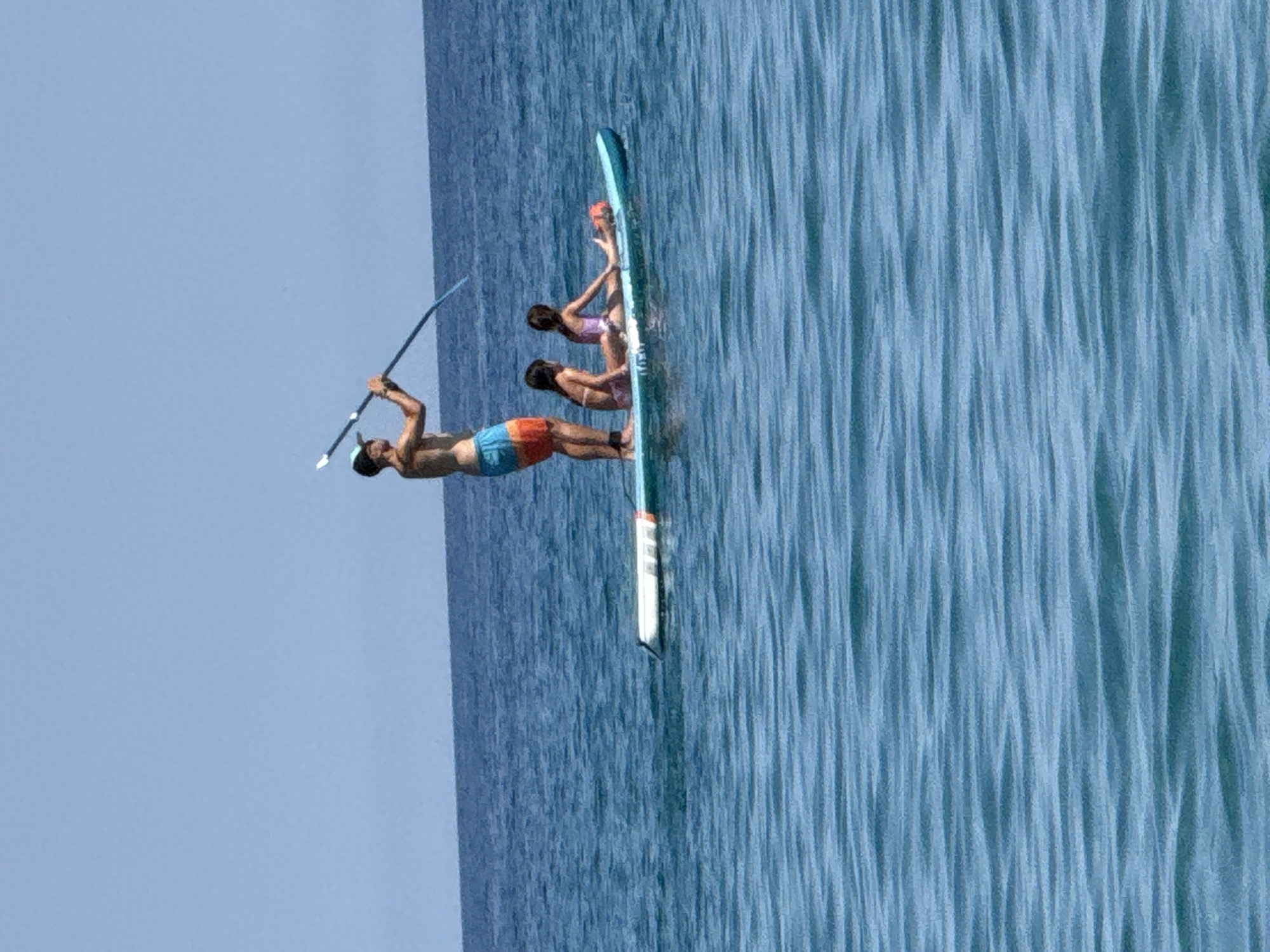 This image depicts three people on a paddleboard in the ocean. One person, a man wearing blue and orange striped swim trunks, is standing on the paddleboard holding a paddle. He appears to be looking towards the left of the frame. Behind him, two other people, seemingly a woman and a child or smaller person, are sitting on the paddleboard. The woman is wearing a pink and purple bikini. The paddleboard is light blue. The water is a vibrant blue with visible ripples and texture, suggesting it is either choppy or the light is creating this effect. The sky is a clear, pale blue. The overall scene suggests a sunny day, likely midday or afternoon given the brightness. The individuals appear to be enjoying a recreational activity. There is no visible text in the image. The location context of Blanes, Spain, implies a coastal setting, likely a beach or open sea area.