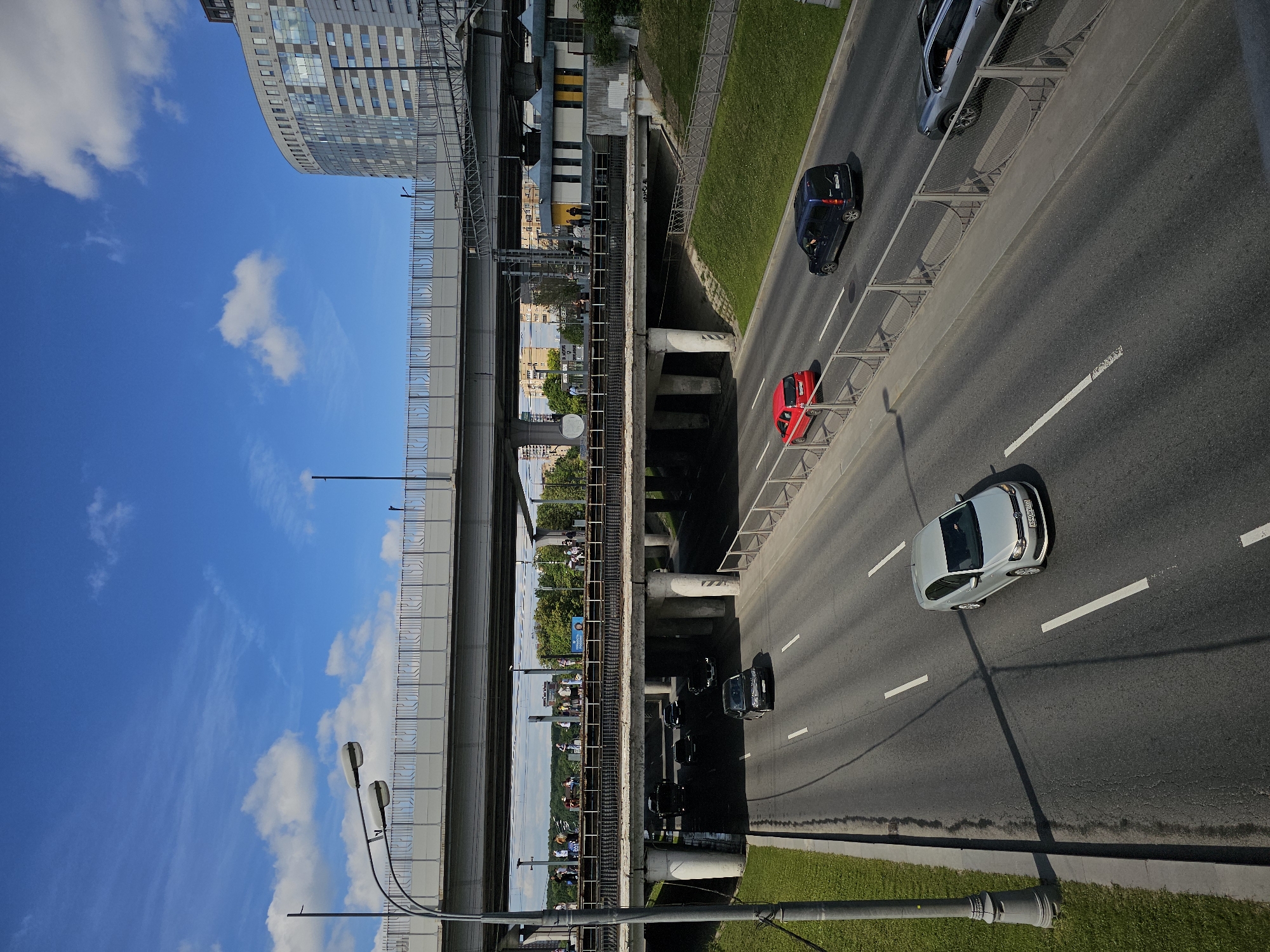 This is an eye-level, high-angle shot of a multi-level urban highway intersection in Moscow, Russia, on a bright, sunny day with scattered clouds. The foreground features a paved road with lane markings, where a silver Volkswagen sedan is driving in the right lane. To the left, a gray lamppost stands on a grassy verge.
Above the foreground road, another level of the highway is visible, supported by concrete pillars. This upper level is populated with several cars, including a red hatchback, a dark blue van, and a dark SUV. More traffic is visible on the furthest upper level, with people walking along a pedestrian overpass.
In the background, a modern, curved glass-fronted building dominates the skyline. Trees and other buildings are visible beneath the highway overpasses, suggesting a dense urban environment. The scene depicts a busy transportation hub in Moscow.