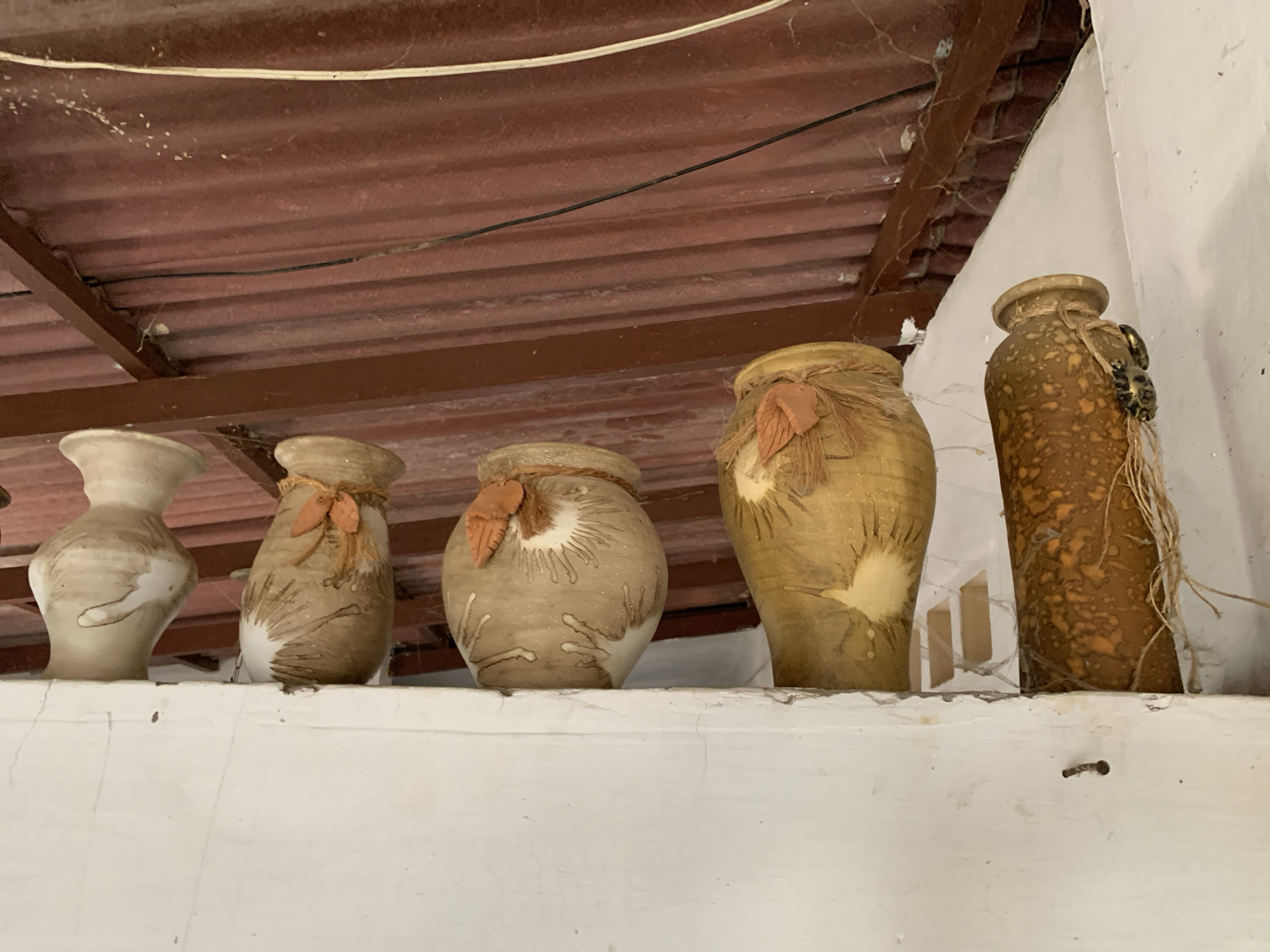 The image displays a still life arrangement of five decorative vases or pots positioned on a white, textured shelf. The pottery, crafted from what appears to be ceramic or clay, exhibits varying shapes, sizes, and earthy color palettes including beige, light brown, dark brown, and yellowish hues. Several vases are adorned with natural twine and small, leaf-shaped ornaments. The vase on the far right features a distinctive dark, lumpy decorative element and loose string hanging from its neck.
Above the shelf, a reddish-brown corrugated metal ceiling structure is visible, crisscrossed by a white and a black wire. Numerous cobwebs cling to the ceiling and its supporting beams, suggesting the space is either aged, rustic, or not frequently maintained. The wall to the immediate right of the display is white. The ambient lighting indicates daylight, but not direct sunlight. The scene depicts an interior or covered outdoor area, possibly in Santa Ana, Venezuela, with a rustic and lived-in aesthetic. No activity is taking place, and there is no visible text.