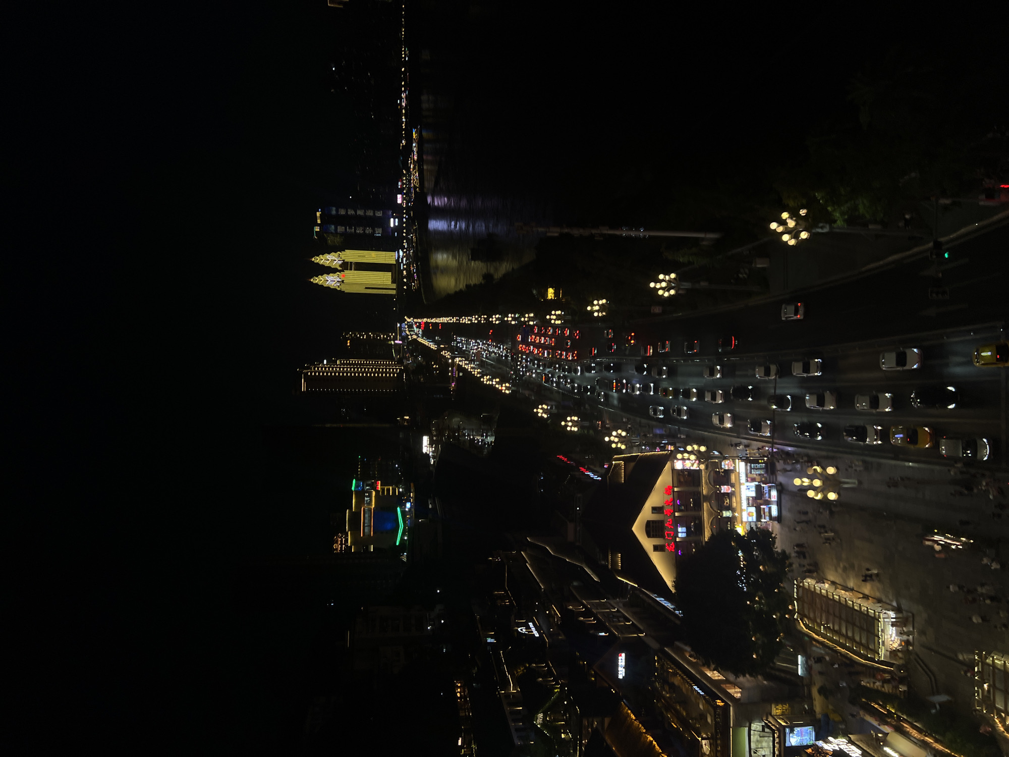 This is a nighttime aerial view of a city street and riverfront in Nan'an District, China. The scene is illuminated by the artificial lights of buildings, streetlights, and vehicle headlights.
On the left side of the image, there are several buildings with illuminated facades, some featuring neon signs. One building has a large white triangular roof with red characters on it. A pedestrian walkway is visible in front of these buildings, with people walking and gathered. A long, covered walkway or structure with internal lighting is also present.
The main focus of the scene is a busy multi-lane street running through the center of the image. Numerous cars, appearing as points of light from headlights and taillights, are moving in both directions, indicating traffic. Streetlights with multiple bulbs line the side of the road.
To the right of the street, a wide river flows, reflecting the lights of the city. Several boats or ships are visible on the water. In the distance, across the river, a cluster of illuminated skyscrapers is visible, with two prominent twin towers lit in yellow.
The overall impression is one of urban activity and vibrant nightlife. The darkness of the night sky contrasts with the bright man-made lights that define the urban landscape.