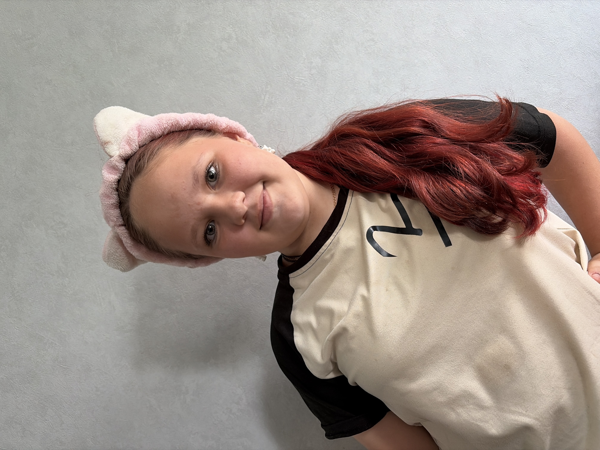 A young person with red hair and a pink headband with ears is wearing a tan t-shirt with black sleeves and a black number "21". The person is smiling and looking at the camera. The background is a light grey wall. The location is unknown and not evident in the provided image.