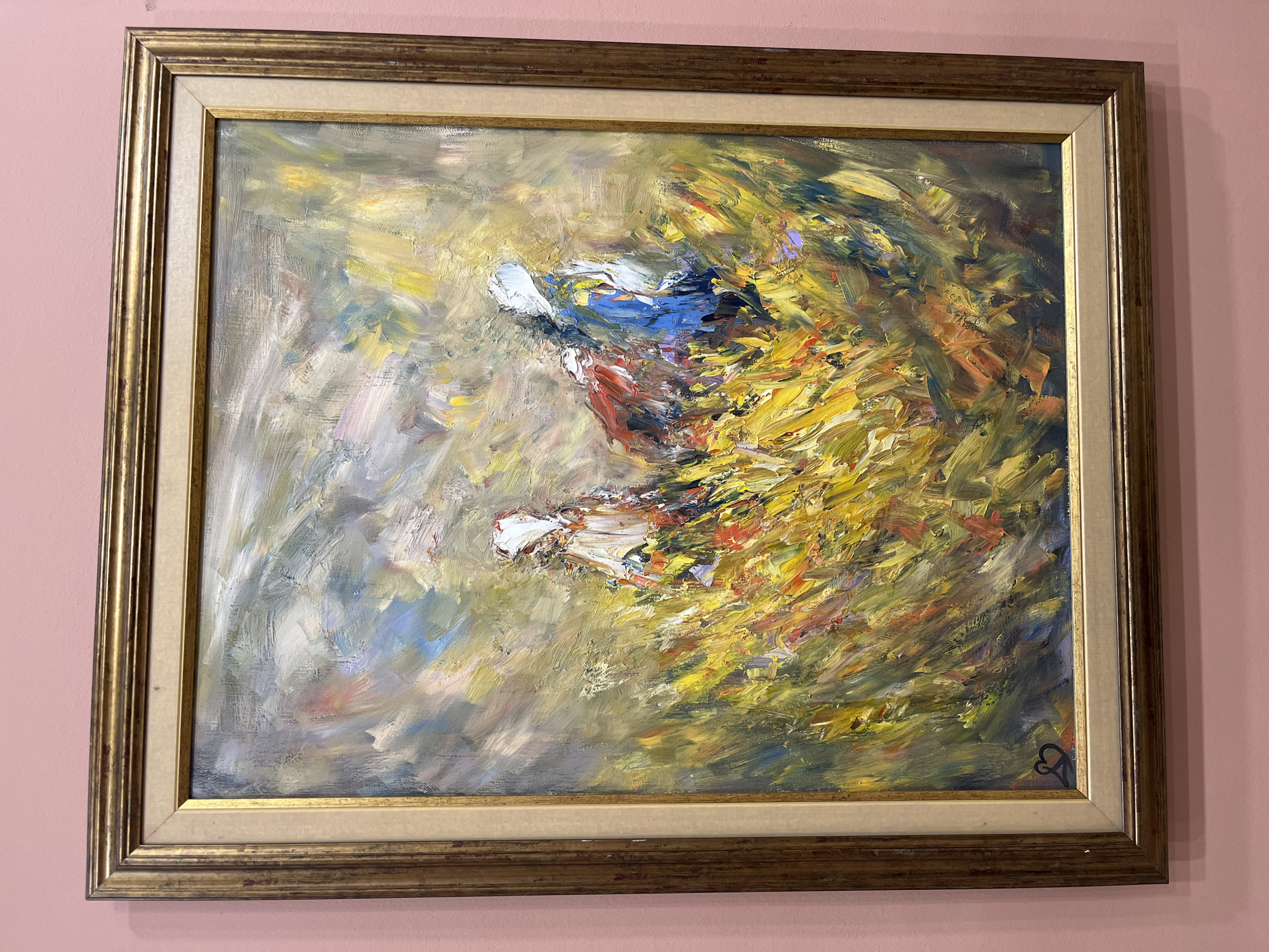 The media file features an abstract oil painting, encased in a gilded frame, hanging on a light pink wall. The painting is characterized by thick, impasto brushstrokes and a dynamic color palette predominantly of vivid yellows and oranges, with significant accents of white, blue, and touches of red and darker tones.
The central and right portions of the canvas depict a swirling, energetic mass of warm colors, suggesting intense motion or a turbulent scene. Within this vibrant activity, more defined, lighter forms in white, blue, and red emerge, which are highly stylized and could be interpreted as figures, wings, or elements of a dynamic creature in action or flight. The left side of the painting presents a softer, more subdued area with cool tones of grey, light blue, and white, providing a contrasting backdrop to the central energy.
The artwork is set within an ornate, gold-colored wooden frame with a decorative inner lip and an additional cream-colored linen or fabric liner. In the bottom right corner of the painted canvas, a distinct black symbol is visible, appearing to be a stylized heart or a monogram. The painting conveys a strong sense of movement, light, and emotional intensity. This media file is located in Plovdiv, Bulgaria.