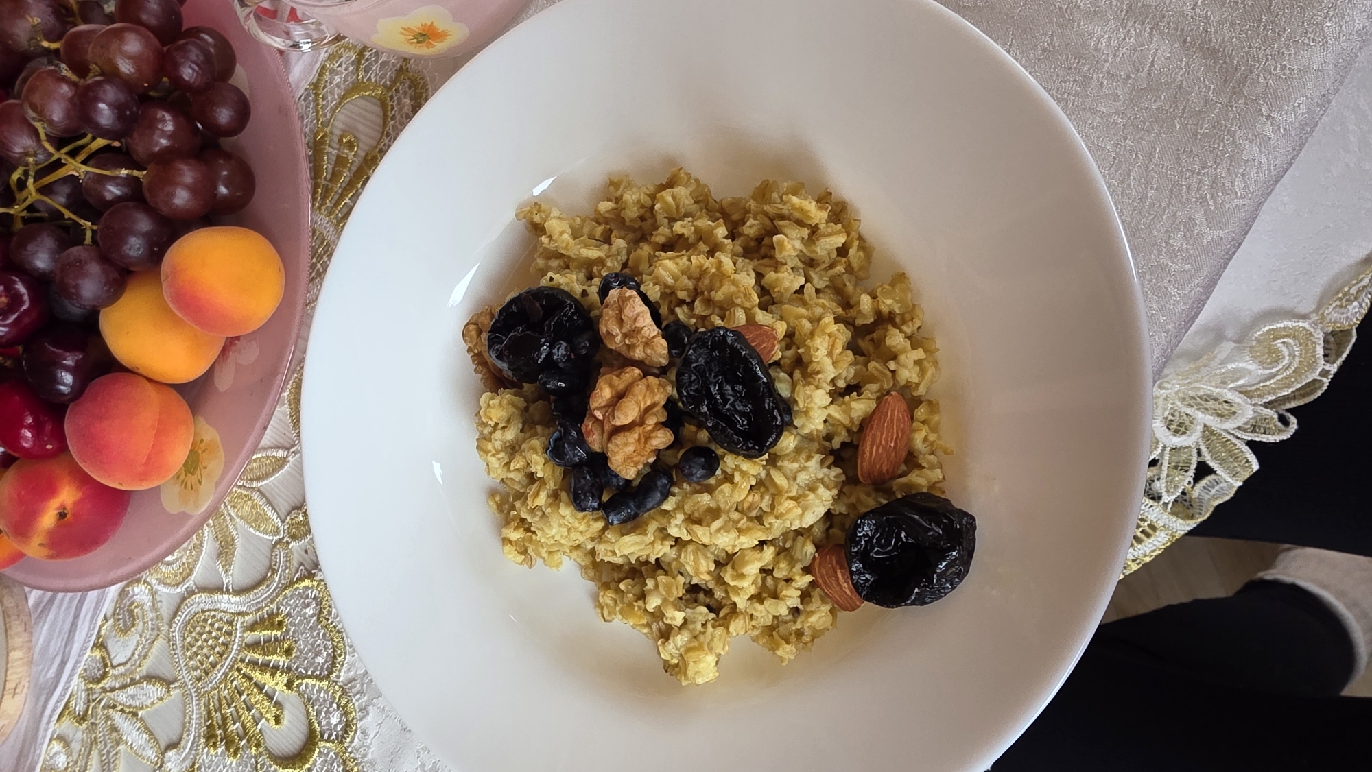 The image shows a white plate filled with oatmeal topped with walnuts, prunes, and blueberries. In the background, a pink plate of fruit, including apricots and grapes, is visible on a table with a decorative cloth. The scene appears to be an indoor setting, possibly a home or a cafe, in the village of Kupoyarovo, Russia.