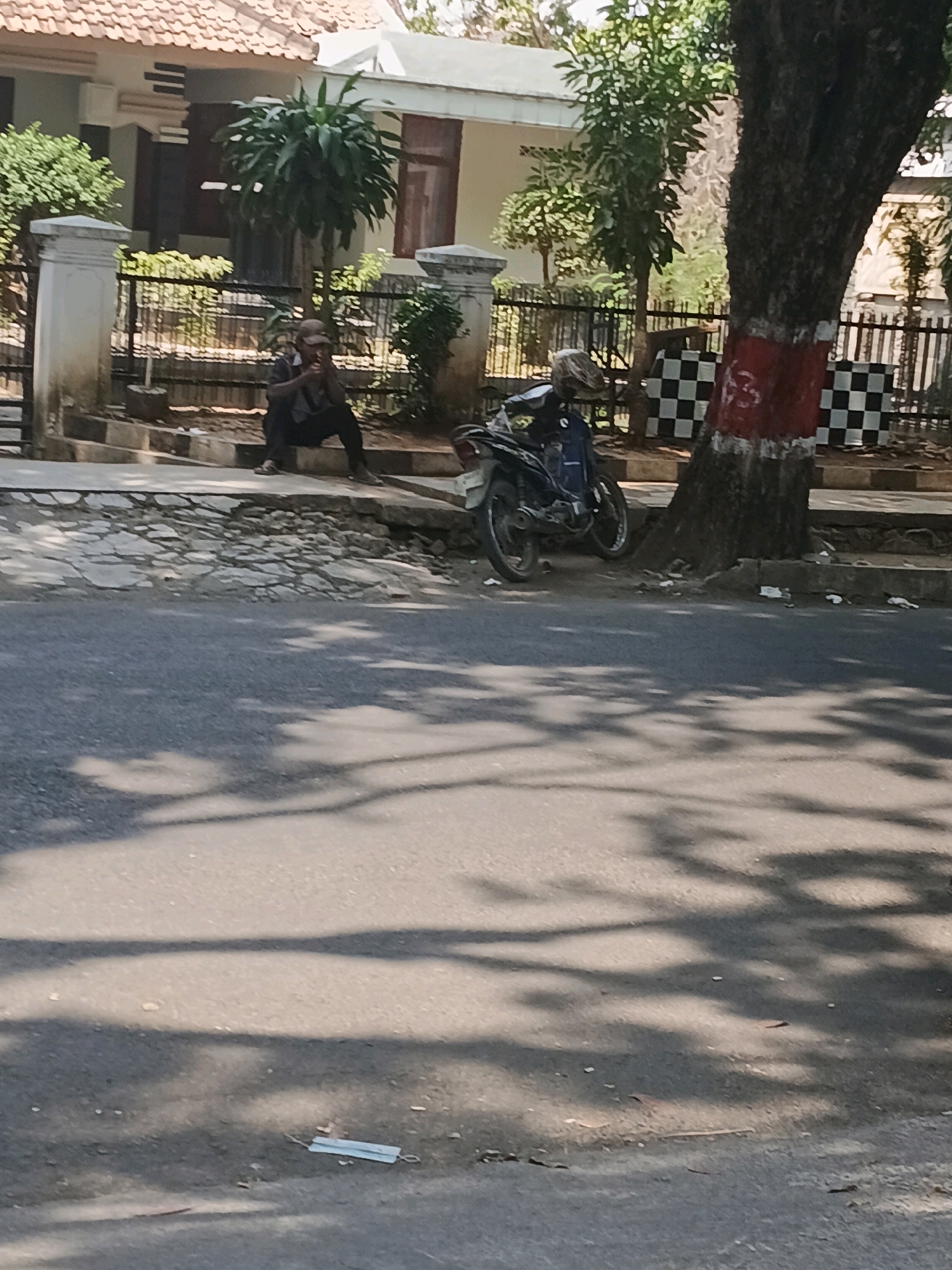 The image shows a man sitting on a low wall by the side of a road in Purwodadi, Indonesia. He is wearing a cap and a dark shirt, and appears to be smoking. Next to him, a motorcycle is parked on the sidewalk. The road is paved and appears to be in moderate condition. A tree with red and white paint markings is located to the right of the motorcycle, and a checkered wall is visible behind it. Buildings with tiled roofs are in the background. The scene is illuminated by sunlight, casting shadows on the road. A discarded face mask is visible on the road surface in the foreground.