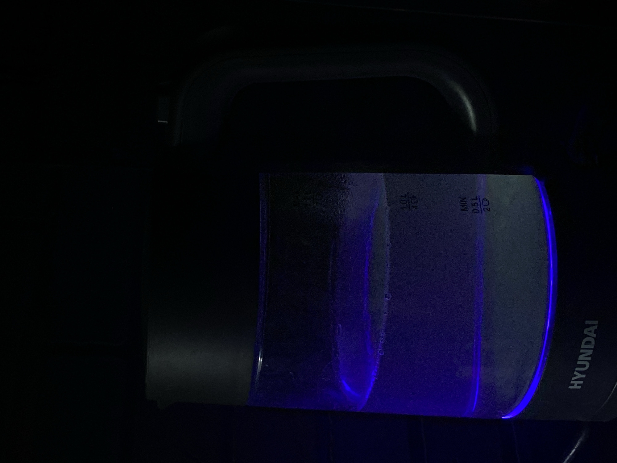 A Hyundai-branded electric kettle filled with water is illuminated by blue light in a dimly lit room. The water level is above the minimum mark. The handle of the kettle is visible. The scene lacks discernible details about the location in Sochi, Russia. No people are visible.