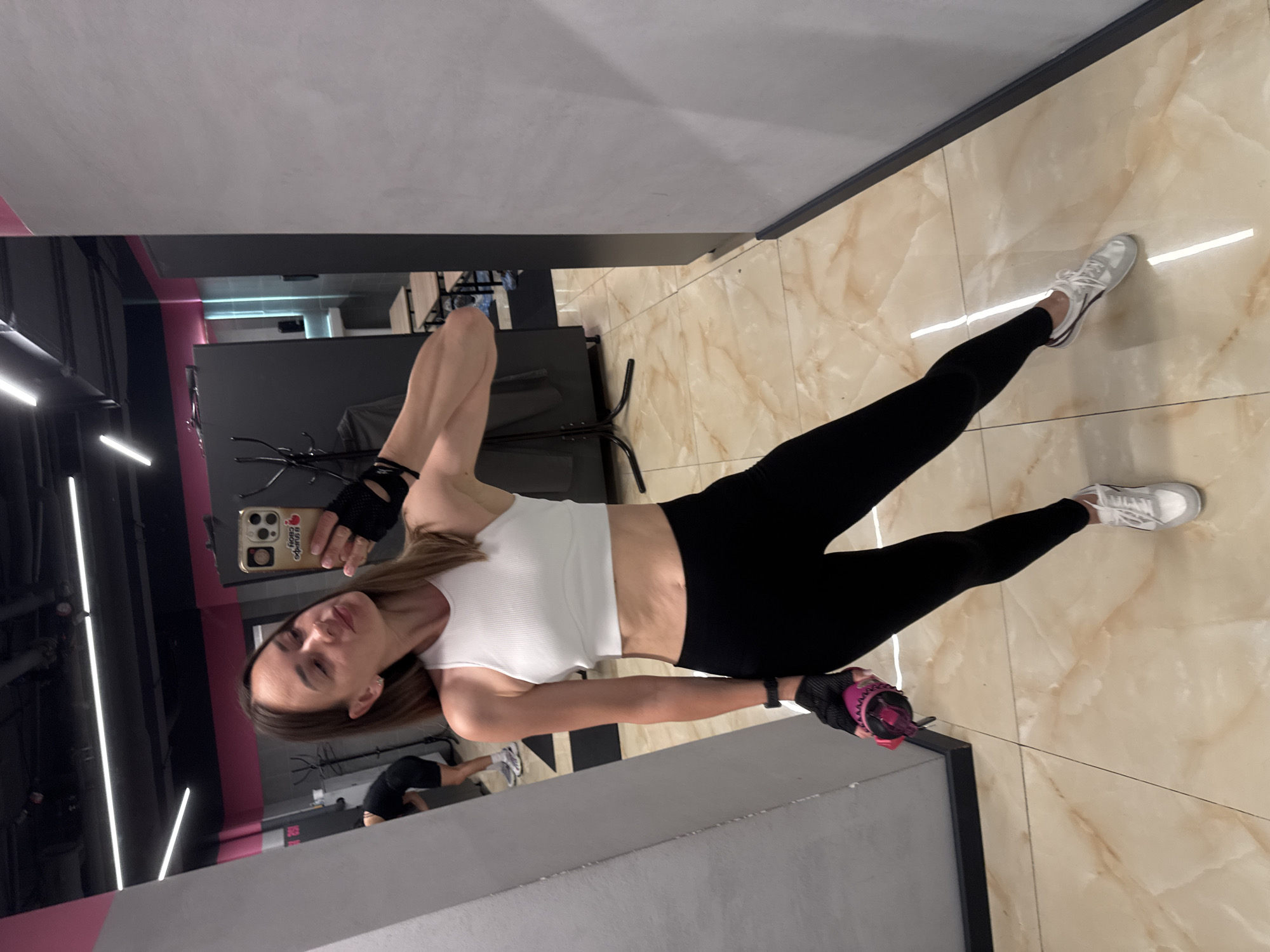A young woman with brown hair is posing for a selfie in a gym. She is wearing a white crop top, black leggings, and white sneakers. She has black fingerless gloves on her hands and is holding a pink water bottle. The gym has a tiled floor and the walls are grey and pink. In the background, there are gym equipment and mirrors. The woman is looking at the camera and smiling. There is text visible on her phone case: "В СВОЁМ РИТМЕ" which translates to "IN YOUR OWN RHYTHM." The image is taken during the day.