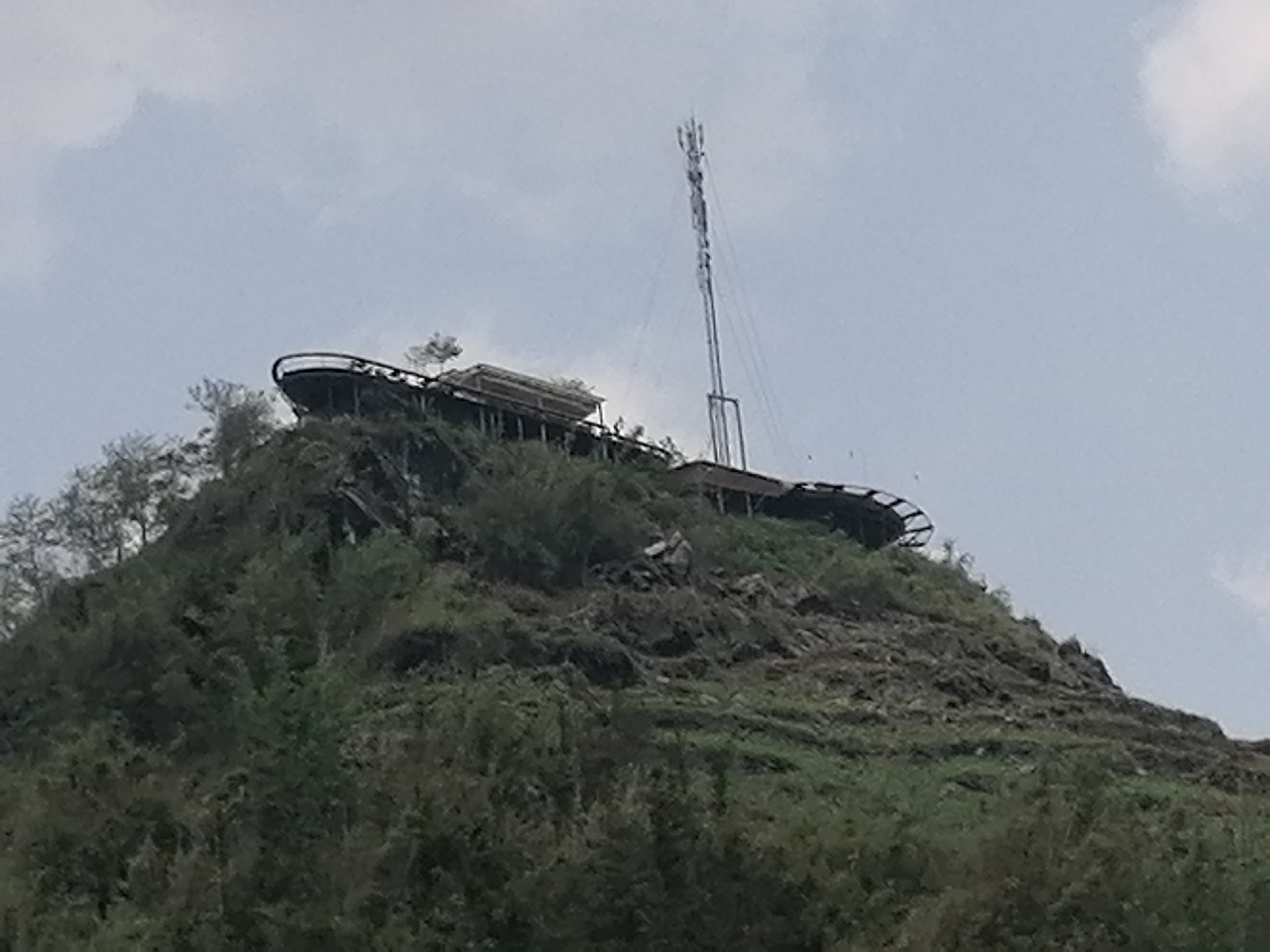 The image displays a steep, verdant hill or mountain, characteristic of the landscape in Sa Pa Ward, Vietnam, under a mostly overcast sky. The hill is densely covered with green trees and thick vegetation, interspersed with rocky sections.
Perched atop the summit is a man-made structure designed as a viewing platform or walkway, featuring a long, curved path with railings that wraps around the peak. A small, rustic building or shelter with a light-colored roof is situated on this platform. Adjacent to the platform, a tall, slender metal communication tower, likely a cell tower or radio mast, stands prominently, extending upwards into the grey sky.
The scene depicts a natural, mountainous setting that has been adapted with tourist infrastructure for observation and a communication facility. The time of day is daytime, with soft light indicating an overcast or cloudy weather. No people are visible, and no specific activity is taking place. There is no visible text in the image.